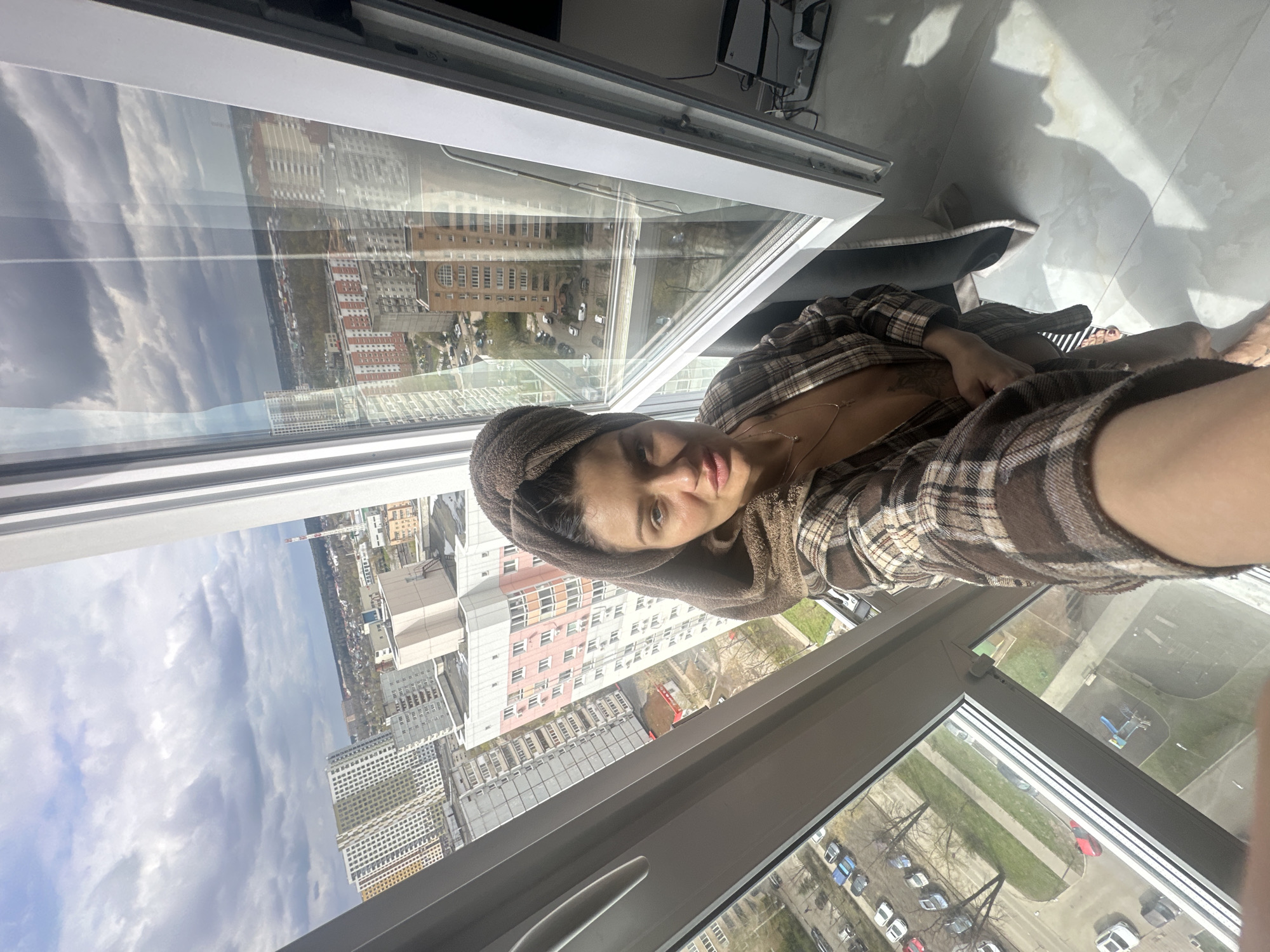 A woman wearing a brown towel wrapped around her head and a plaid robe is photographed from a high-angle perspective indoors, near a window. The window offers a view of an urban landscape, suggesting she is in a tall building. Outside, one can see buildings of various sizes and colors, including both high-rise apartment blocks and lower-rise structures, interspersed with green spaces and streets filled with cars. The sky is overcast with clouds. The location is Moscow, Russia. The interior setting appears to be a domestic space, likely her apartment, as indicated by the personal items visible in the reflection of the window.