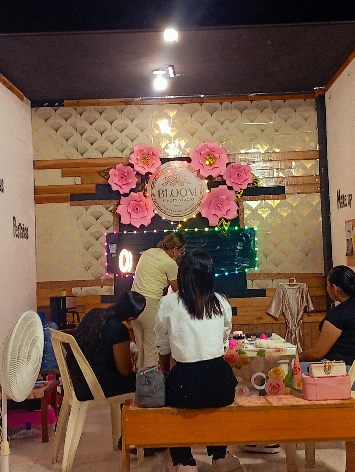 The image shows the interior of a beauty studio in Porlamar, Venezuela. Three women are present, two seated and one standing. One seated woman is facing away from the camera, wearing a white shirt and black skirt. Another seated woman is facing to the right, wearing a black top. A third woman is standing, wearing a beige top and pants, possibly providing a service to one of the seated women.
The background features a wall decorated with a "Bloom Beauty Studio" logo surrounded by large pink paper flowers. There's also a neon ring light and colored string lights. The walls are partially covered with decorative paneling and wallpaper.
Additional objects include a fan, water dispenser, table with cosmetic items, a garment hanging on the wall, and various small storage boxes. The room appears to be a small, enclosed space, likely a storefront or a section within a larger building.