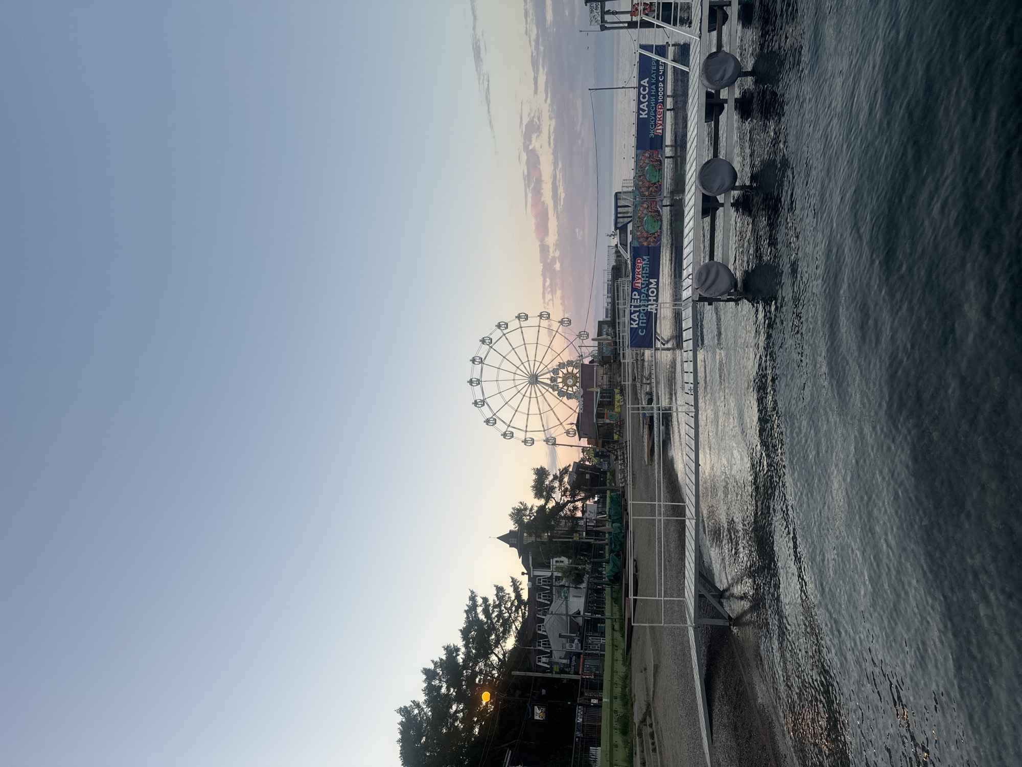 A wide shot captures a twilight scene in Listvyanka, Russia, overlooking Lake Baikal. In the foreground, a wooden pier extends into the dark, rippling water. Three large, round objects, covered with gray tarps, are secured to the pier.
Further back, a sandy beach leads to a promenade lined with trees and buildings. A Ferris wheel is prominently displayed, its structure illuminated against the soft pastel hues of the sunset sky. Signs are visible on the promenade, one reading "КАССА" (KASSA, meaning "TICKETS" in Russian) and another with text related to boat excursions. The overall atmosphere is calm and serene, with the natural beauty of the landscape contrasting with the man-made attractions.