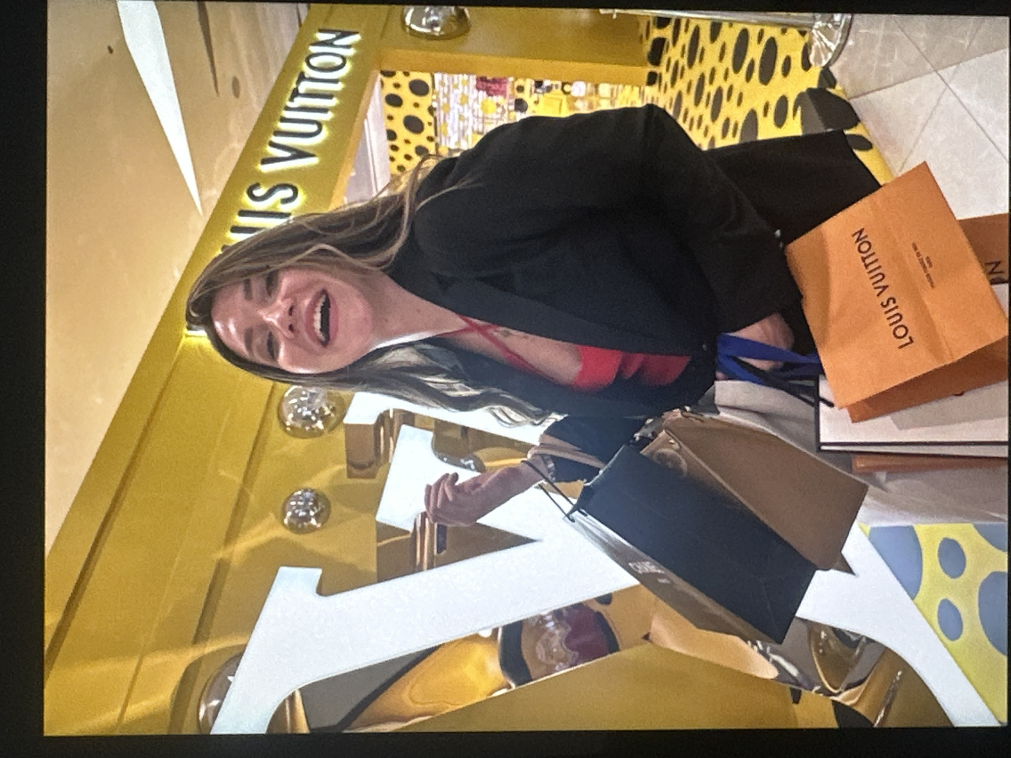The image shows a woman standing in front of a Louis Vuitton store with two shopping bags, one Louis Vuitton and the other Chanel. She is wearing a black blazer and red top. The interior of the store is visible, featuring a yellow and black polka dot pattern. The location appears to be within a city, but the specific city and state are unknown based on the image.