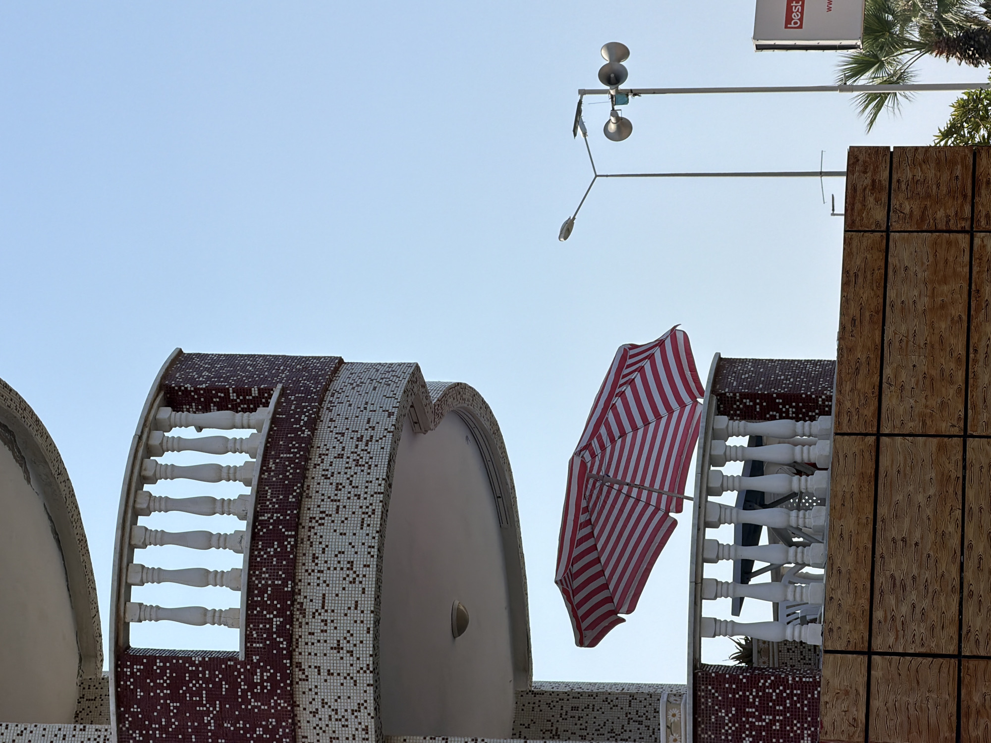A balcony of a building in Alanya, Turkey, is visible against a clear blue sky. The balcony features a white balustrade and is partially covered by a red and white striped umbrella. A table and chairs are visible on the balcony. In the background, there is a lamppost with lights and loudspeakers, and a billboard with the word "best" is partially visible. The lower part of the image shows a wall with a wood-grain texture. The overall scene suggests a sunny day.