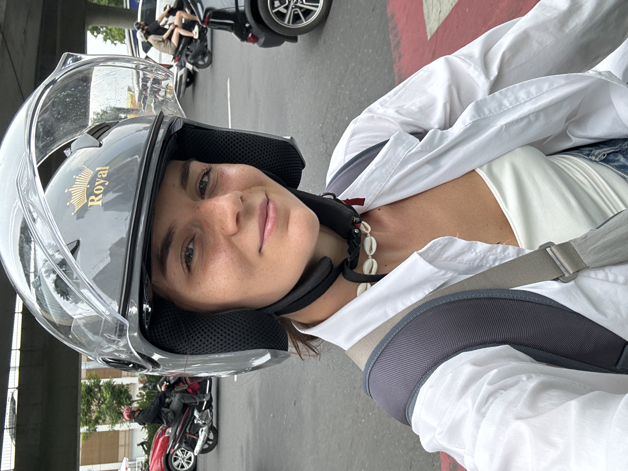 A woman wearing a gray helmet with a clear visor, labeled "Royal", and a white shirt with a gray backpack strap over her shoulder, is likely taking a selfie. She is facing the camera with a slight smile. Around her neck is a necklace made of shells.
In the background, there are scooters and a red and white painted line on the road. There are other people riding scooters in the distance and a building to the left.
The image was likely taken on a street in Thủ Đức, Vietnam, as evidenced by the scooters and general urban environment.
