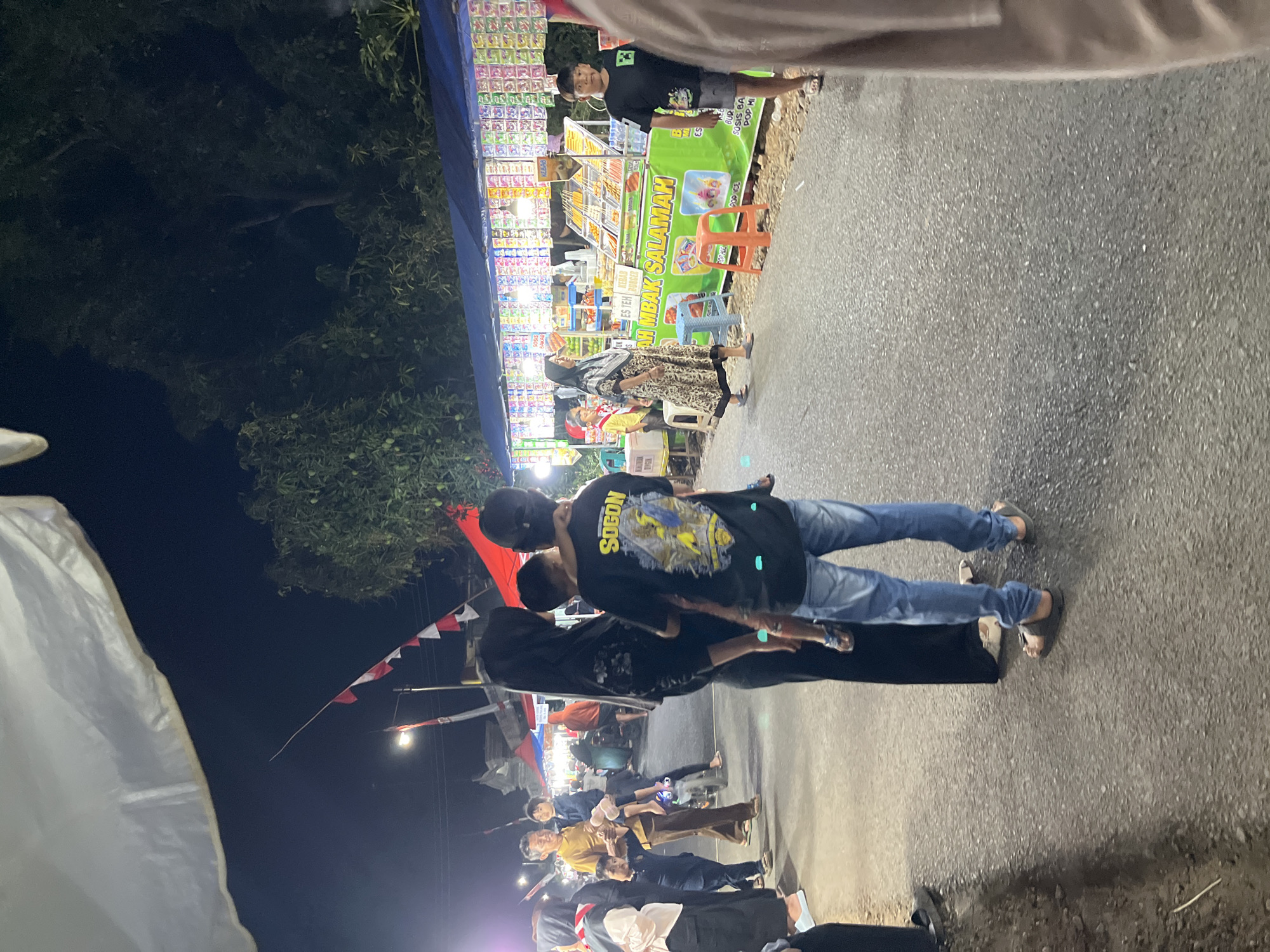 The image depicts a nighttime street scene in Gembong, Indonesia, likely at a market or fair. A group of people are visible, some carrying children. On the right side of the image, a food stall is set up with various items displayed, including what appears to be snacks and drinks. The stall is brightly lit, illuminating the products. Flags, possibly Indonesian flags, are strung across the street in the background. Trees and foliage are visible behind the stall, and a dark sky suggests it is nighttime. The ground is paved, indicating a road or a street. The overall atmosphere appears to be lively and social, with people browsing the stalls and interacting.