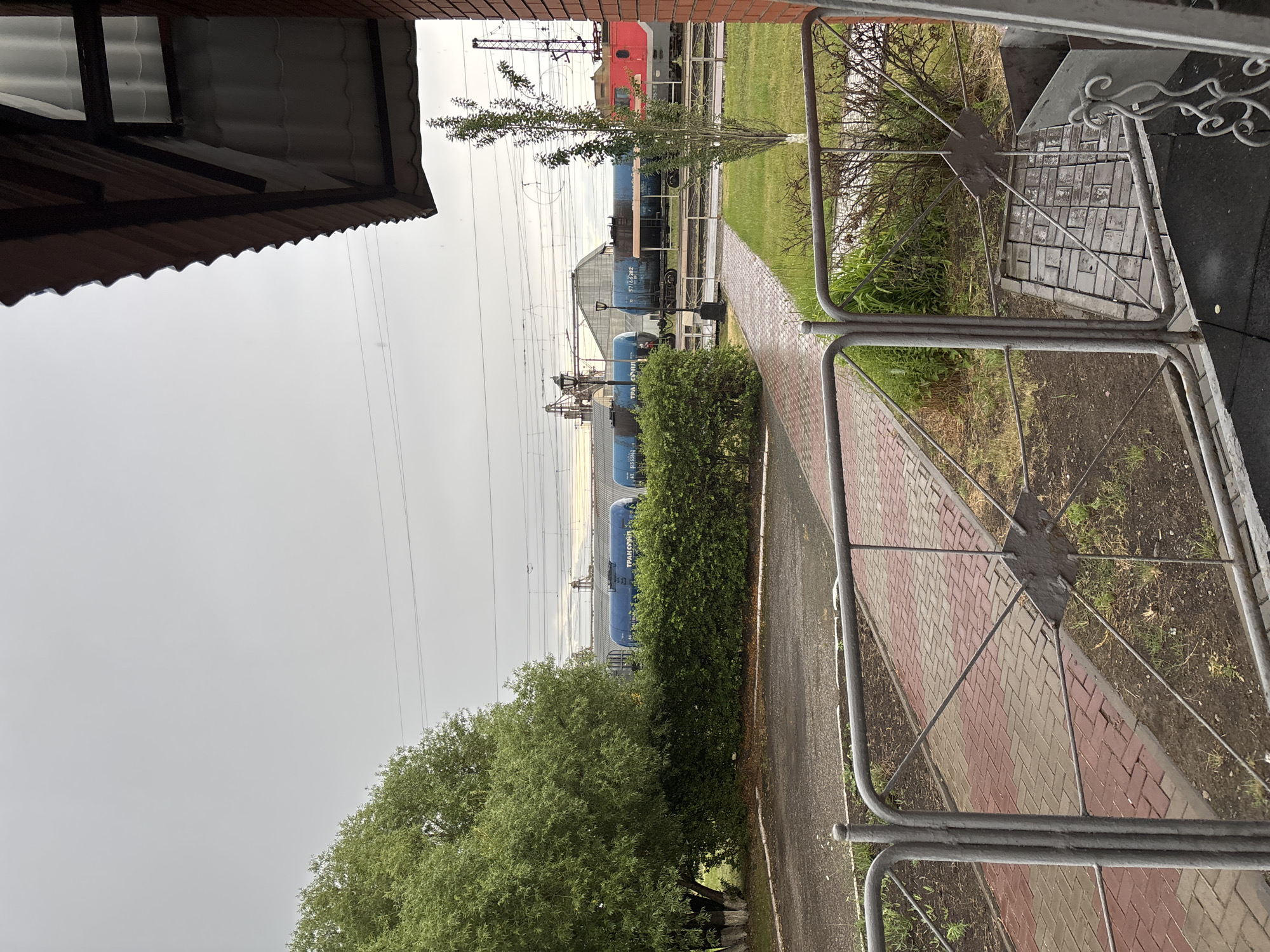 An overcast, possibly rainy day at a train station in Moskalenki, Russia. The foreground is dominated by a metal fence. Beyond the fence, a red brick building's roof and wall are visible on the right. A paved walkway, laid with red and beige bricks, leads away from the viewer and curves to the left. To the left of the walkway is a patch of dry, dark soil with some scattered grass. Further left, a large, lush green tree with a white-painted base stands near a neatly trimmed green hedge. Behind the hedge, a row of blue train cars with "ТРАНСОЙЛ" (Transoil) written on them are parked on train tracks. In the distance, more train tracks and overhead power lines are visible, along with industrial buildings and a red locomotive on the far right. The sky is a uniform gray.