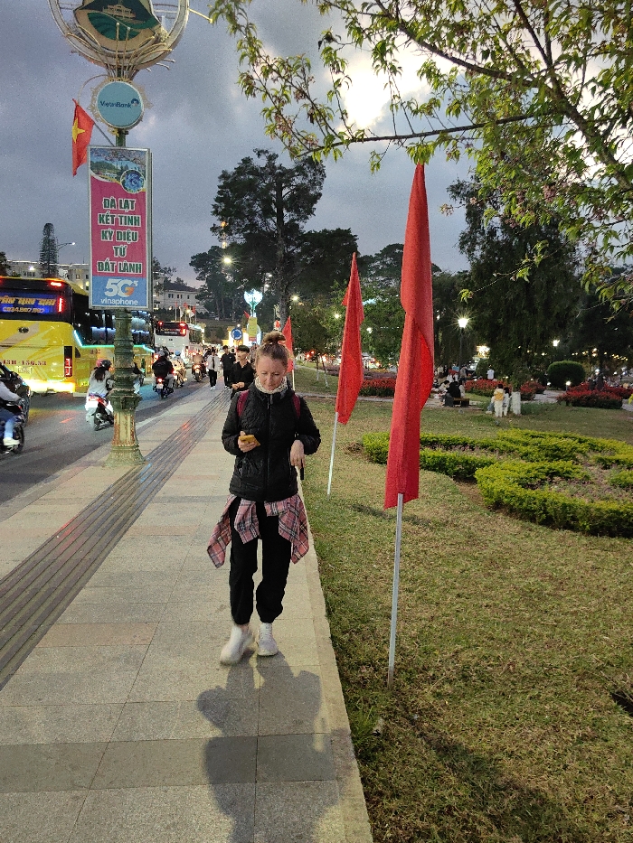 The image depicts an urban street scene in Da Lat, Vietnam, during dusk or early evening. The specific location is identifiable by a tall lamppost on the left, adorned with a Vietnamese flag and a vertical banner displaying "ĐÀ LẠT KẾT TINH KỲ ĐIỆU TỪ ĐẤT LÀNH," along with "Vietcombank" and "Vinaphone 5G" logos.
In the foreground, a Caucasian female individual, approximately 20-30 years old, walks on a light-colored paved sidewalk. She is dressed in a black quilted jacket, black sweatpants, white athletic shoes, and has a red and white plaid shirt tied around her waist. Her hair is pulled into a bun. She is holding a yellow smartphone in her right hand and appears to be looking down at it. Her shadow is cast behind her on the pavement.
To the right of the sidewalk, a grassy park area features two prominent, solid red flags affixed to slender metal poles. The park includes manicured green bushes and visible red flowers in beds. Further into the park, several individuals are visible, some seated, amidst trees and illuminated decorative lights.
To the left of the sidewalk, a busy street is active with traffic. A large yellow passenger bus with "TÂN THI" partially visible on its side is prominent. Numerous motorcycles and scooters, ridden by individuals wearing helmets, are also present, along with other vehicles. Tall trees line both sides of the street and park, extending into the background. The sky above is overcast, and multiple streetlights are illuminated throughout the scene.