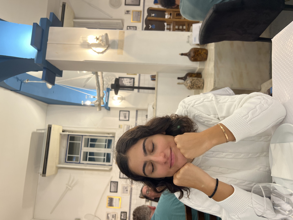 The image shows the interior of a restaurant in Coruche, Portugal. A female individual is positioned centrally in the foreground, seated at a table. She has dark, wavy, shoulder-length hair, her eyes are closed, and a faint smile is present. Her hands are clasped together, resting beneath her chin. She wears a white cable-knit sweater, a black beaded bracelet on her left wrist, and a thin gold-colored bangle on her right wrist. On the table directly in front of her, a white placemat, a folded white napkin, and an empty clear glass are visible.
The background reveals the restaurant's decor, characterized by white walls adorned with various framed pictures and a decorative white relief of a fork and spoon. A blue and white tiled dado rail runs along the lower section of the walls. An air conditioning unit is mounted above a white and blue-trimmed interior window frame. The ceiling features a prominent light blue arched section and structural elements. A large, nautical-themed light fixture, composed of light-colored branches with integrated lantern-style lights, hangs from the ceiling. Additional lantern-style wall lights are affixed to pillars and walls throughout the space. In the mid-ground, a counter displays several large, antique-style glass bottles, some containing preserved food items. Other patrons are partially visible in the background; a man in a teal shirt is to the left, and another person in a similar shirt is to the right, seated at separate tables. The lighting within the restaurant is warm.