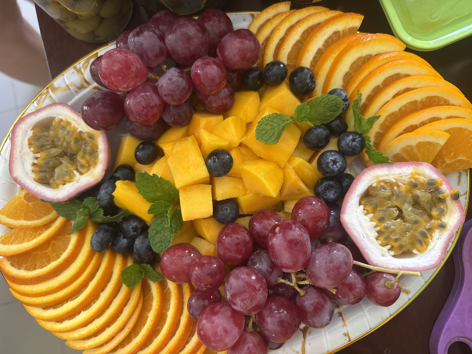 A platter of fruit consisting of orange slices, red grapes, mango chunks, blueberries, passion fruit, and mint leaves. The location is Kuta Selatan, Indonesia.