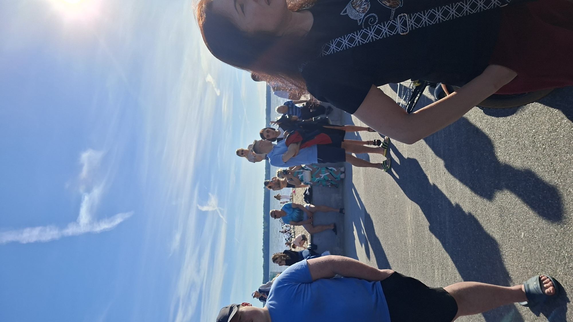 The image captures a group of people outdoors on a sunny day, likely in Tallinn, Estonia. The sun is bright, suggesting it is daytime, and the sky is a vibrant blue with scattered wispy clouds and a visible contrail. The scene appears to be a waterfront or promenade area, with a paved walkway in the foreground and a body of water and a distant shoreline visible.
Several individuals are the primary subjects. In the foreground, a person wearing a blue t-shirt and black shorts is seen from the waist down, with a dark baseball cap. Further back, a woman with dark hair is partially visible in the upper right, wearing a black t-shirt with a patterned design.
The people in the middle ground are standing in a line or small groups, facing towards the water or the distant shore. They are dressed casually in summer attire, including t-shirts, shorts, and sandals, suggesting a warm and pleasant climate. Some appear to be observing something, perhaps a view or an event. One person is wearing a blue t-shirt with a tie-dye pattern and shorts. Another is in a blue t-shirt and patterned shorts. There are bicycles visible in the background, hinting at recreational activities.
The overall mood seems relaxed and leisurely, consistent with a day spent outdoors at a popular spot. The sunlight casts long shadows on the pavement, indicating the angle of the sun. No explicit text is visible in the image.