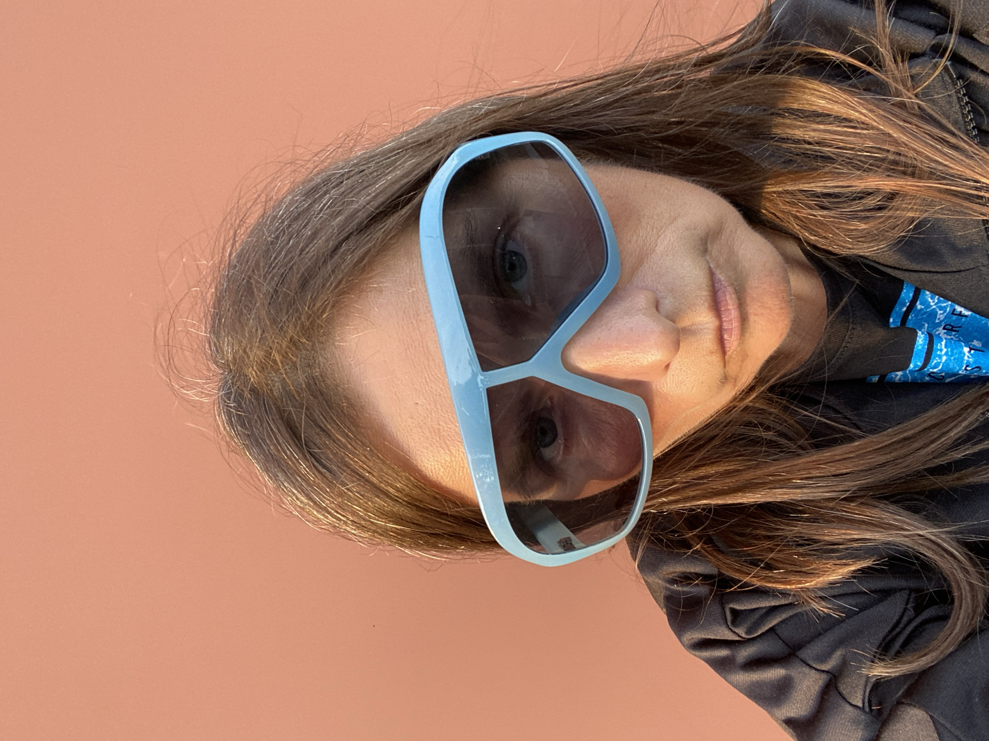 A woman is pictured from the chest up, looking directly at the camera with a serious expression. She is wearing large, light blue wrap-around sunglasses that obscure her eyebrows and part of her eyes. Her dark brown hair is long and worn down, with some strands falling across her face and shoulders. She is wearing a dark jacket over a dark t-shirt with a blue graphic design. The background is a solid, pale orange wall. The lighting suggests it is late afternoon or early morning, with the sun casting a warm glow on the right side of her face and hair. The image appears to be a selfie, taken outdoors against an urban backdrop.