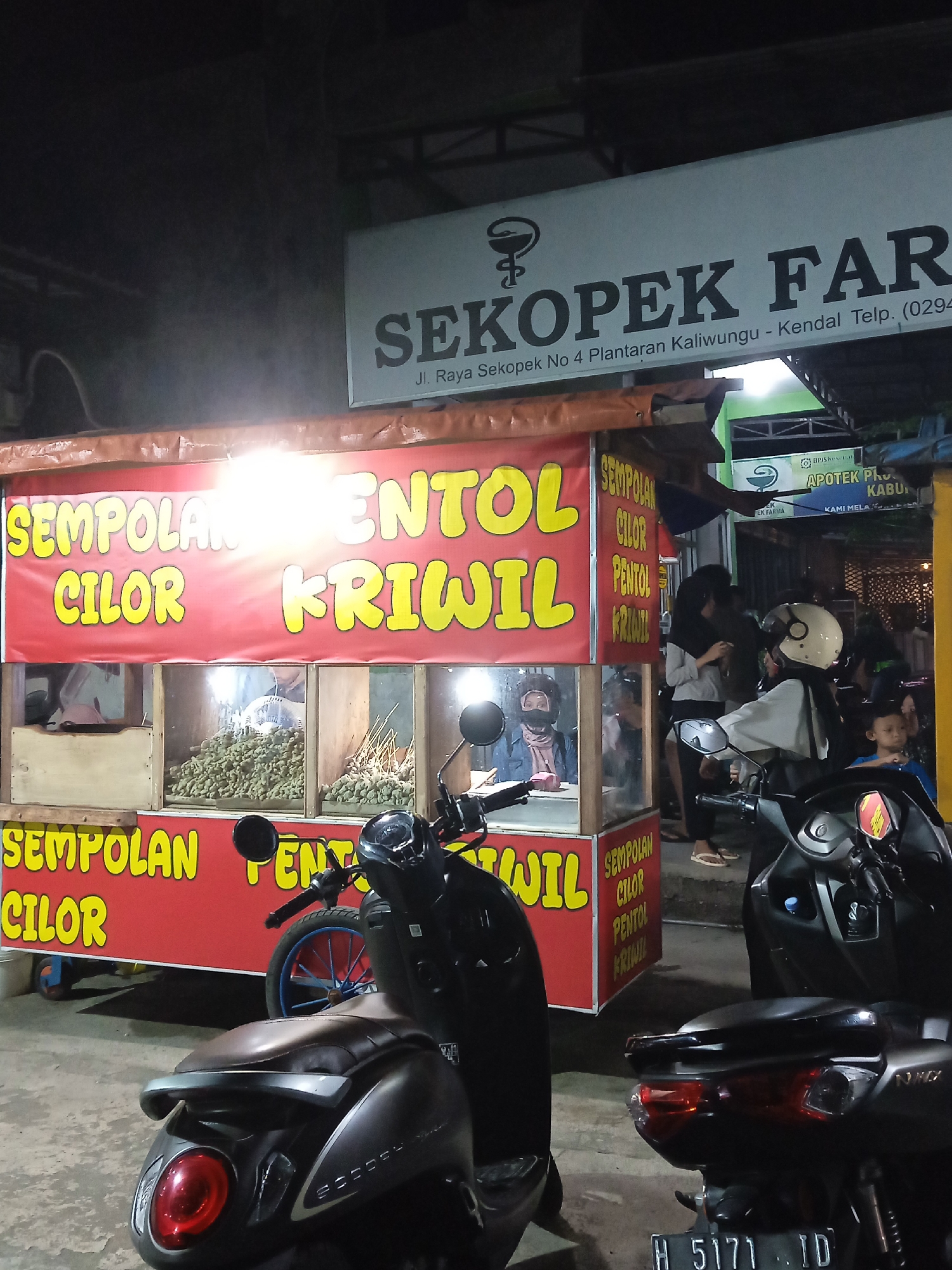 The image captures a night scene in Kebonsari, Indonesia, showing a busy food stall set up alongside a street, with a pharmacy in the background. The sky is dark, and the area is illuminated by artificial lights from the food cart and surrounding establishments.

In the center, a vibrant red and yellow food cart prominently displays various Indonesian snack names. The top banner reads "SEMPOLAN", "CILOR", "PENTOL", and "KRIWIL", with these words repeated on the lower panel of the cart. Inside the cart, behind glass panels, piles of small, skewered snacks are visible, likely the 'pentol' or 'cilok' being sold.

Several people are gathered around the food stall. One person in a light-colored top and dark hijab, wearing a white helmet, appears to be interacting with the stall owner or waiting for an order. Another person with a dark hijab and mask is visible inside the stall. A child can be seen to the right, near other adults.

Multiple motorcycles are parked in the foreground and near the stall, indicating customers. A dark brown scooter is prominent on the left, and a black NMAX motorcycle, with license plate "H 5171 ID", is on the right.

In the background, a pharmacy sign is visible, displaying "SEKOPEK FARMA" and the address "Jl. Raya Sekopek No 4 Plantaran Kaliwungu - Kendal Telp. (0294)". Further details on the pharmacy sign include "BPJS Ke", "APOTEK PH", "KABU", and "KAMI MELA". The overall atmosphere suggests a casual evening street food experience.