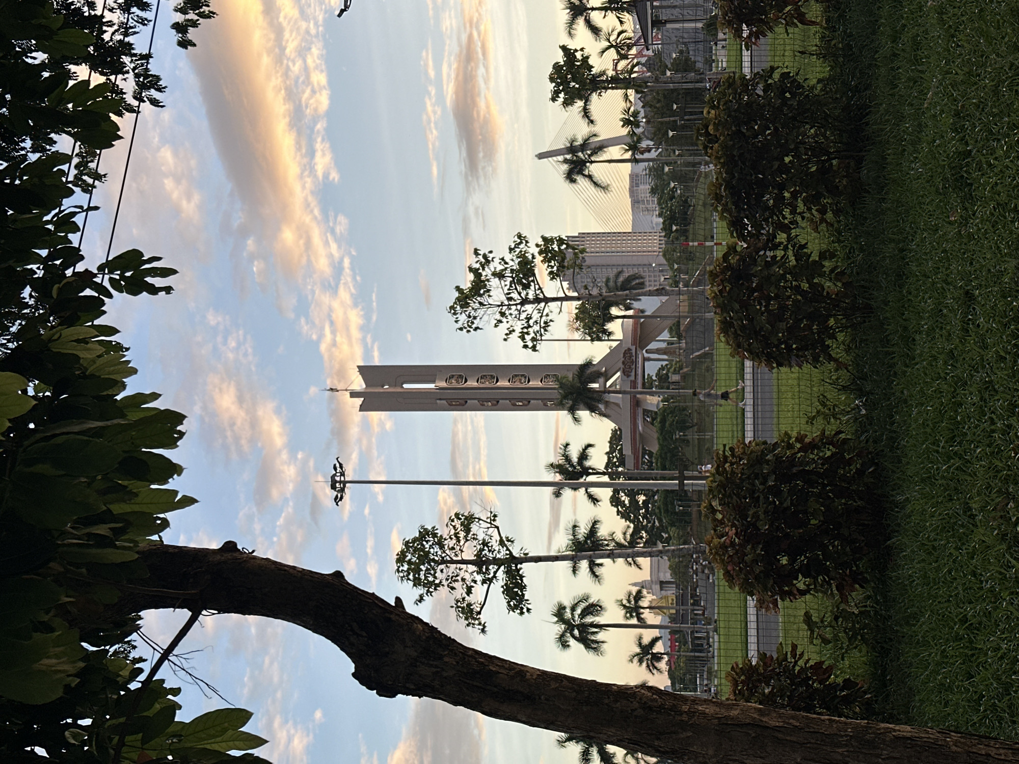 The image depicts a cityscape of Da Nang, Vietnam, at dusk. In the foreground, a large tree trunk occupies the left side of the frame, with its branches and leaves partially obscuring the sky. Behind the tree, a green lawn leads to a row of manicured bushes. A metal fence separates the foreground from a street and a larger park area.
In the mid-ground, a prominent monument stands tall, a concrete structure with geometric shapes and circular elements. Palm trees are scattered throughout the park, some reaching towards the sky. Streetlights are visible, as are buildings in the background. A suspension bridge with its cables is discernible in the distance, suggesting the presence of a river or bay. The sky above is a canvas of pastel hues, with clouds tinged with orange and pink from the setting sun. A few people are visible walking on the street in the distance, appearing as small silhouettes. The overall scene conveys a sense of tranquility and urban beauty as day transitions into night.