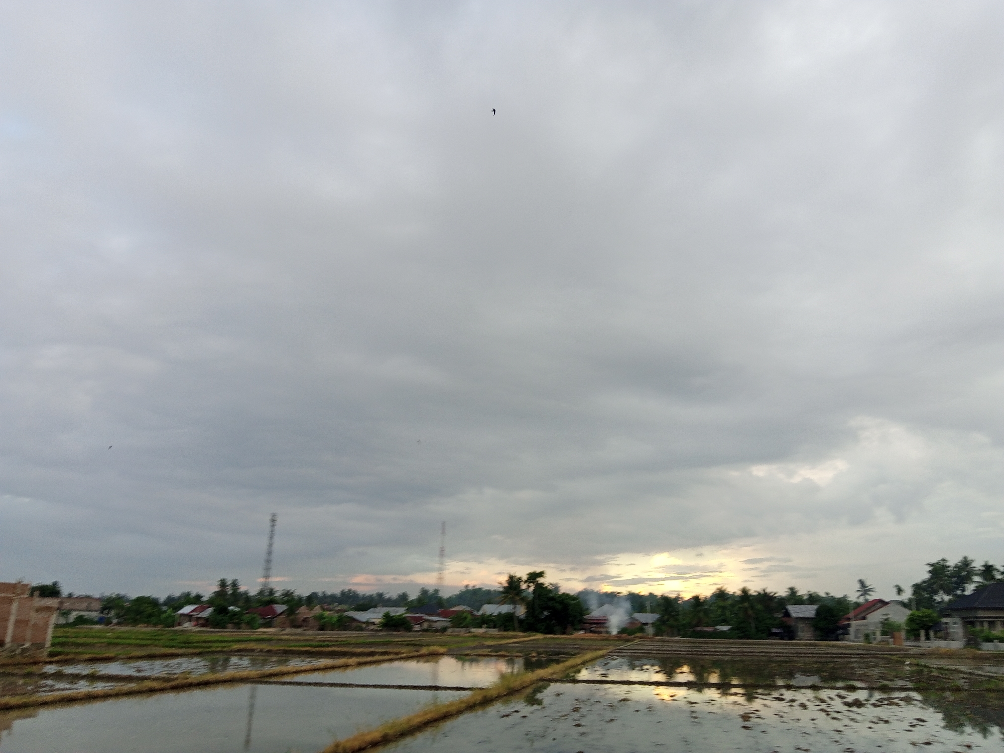 The image shows a rural scene in Geulangganggampong, Indonesia. The foreground is dominated by flooded rice paddies reflecting the sky. In the middle ground are houses and trees, with two tall communication towers visible. Smoke is rising from what appears to be a fire. The sky is overcast with various layers of clouds. There are no visible people or interactions.