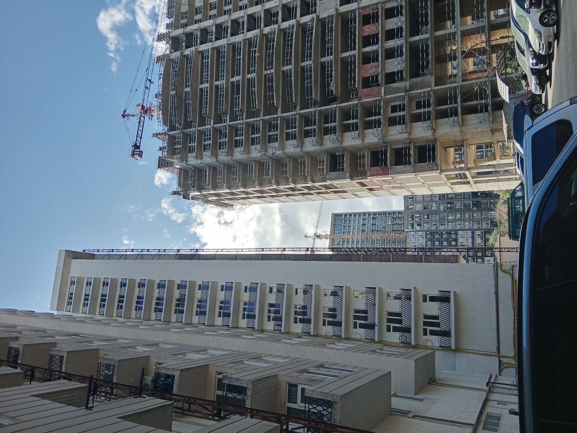 The image captures an upward view in what appears to be a dense urban setting, likely in Batumi, Georgia. Multiple buildings are visible, including one under construction with a crane extending from its top. The buildings vary in height and architectural style. One building is partially obscured by a blue van or car on the right side of the image. The sky is visible with a mix of blue patches and scattered clouds. The photo shows a juxtaposition of old and new construction in the city, with signs of modern development against older structures.