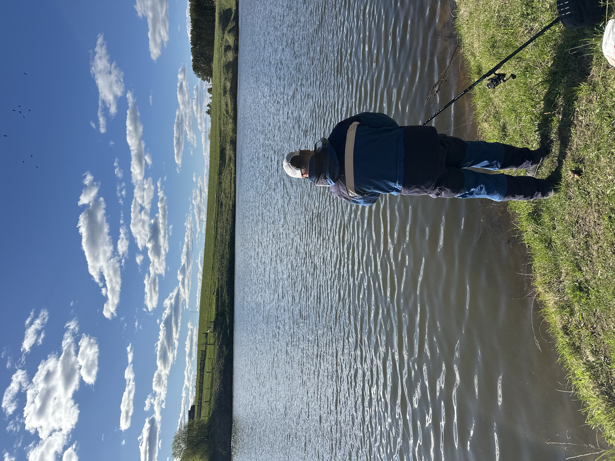 The image depicts a person fishing at a body of water, potentially a lake or pond. The person is wearing a blue jacket with a dark section and a white hat. They stand at the edge of the water, with a fishing rod resting on the grassy bank. The water reflects the sky, which is blue with scattered white clouds. A grassy field and a line of trees are visible in the background. The image may be taken in or near the city of Среднее Кечево, Russia.