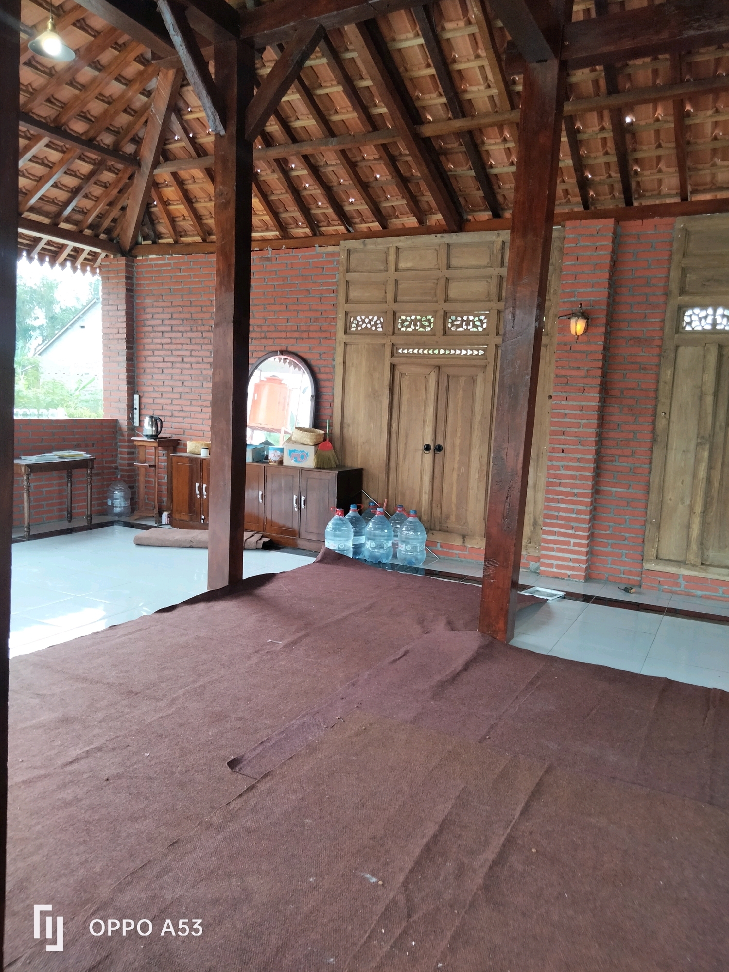 The image depicts an outdoor covered area with a traditional Indonesian aesthetic, likely located in Trenggalek, Indonesia. The structure features a wooden post and beam roof with terracotta tiles. The walls are constructed from red brick and wooden panels. In the background, there are large wooden doors with decorative carvings and a small lamp.
To the left, a wooden table holds a kettle and some papers. A large water jug is on the floor. Adjacent to it is a wooden cabinet with drawers and doors. In front of the cabinet, a large oval mirror reflects a portion of the room, including another large water jug. A broom and dustpan are also visible on top of the cabinet.
In the foreground, a brown carpet covers the floor. Several large plastic water jugs are placed in a row along the wall. The floor appears to be tiled. The overall impression is of a functional, open-air gathering space with elements of rustic charm. The lighting suggests it is daytime.