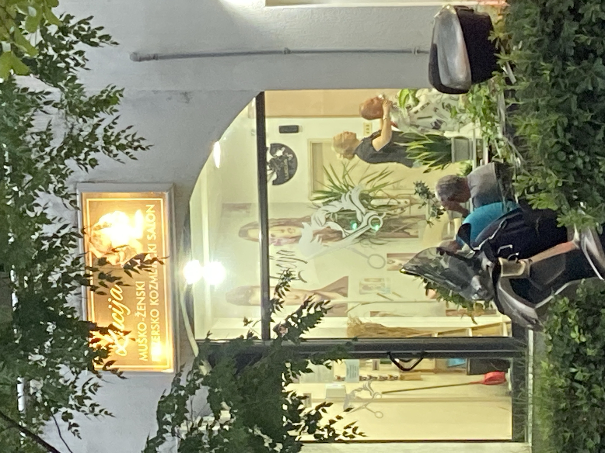 The image shows the exterior of a salon called "Alicija" in Budva, Montenegro. The salon sign reads "Alicija, Muško-Ženski frizersko kozmetički salon." Through the window, a mannequin head is visible, and a person appears to be attending to it. A man wearing a blue shirt, and a backpack is standing outside the salon. A scooter is nearby, parked on the sidewalk. Green foliage partially obscures the view of the salon sign and the scooter. The image is taken at dusk or evening.