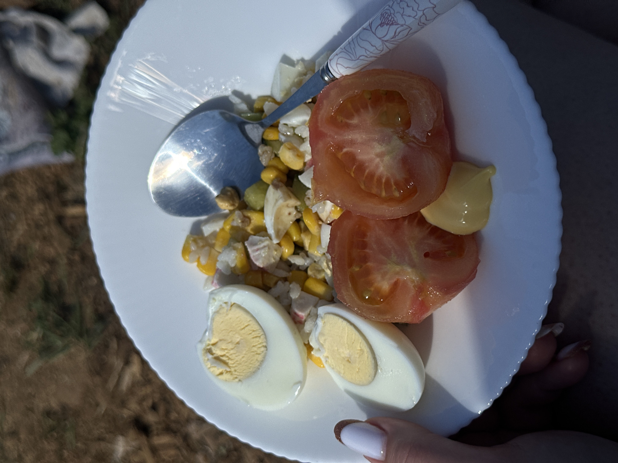 The image depicts a white plate held by a person with manicured nails. The plate contains a salad consisting of corn, chopped egg, surimi, halved tomatoes, and a dollop of mayonnaise. A spoon with a floral design on the handle rests on the salad. The background includes grass, brown soil, and a folded grey blanket. The location is presumed to be in Черёмушки, Russia.