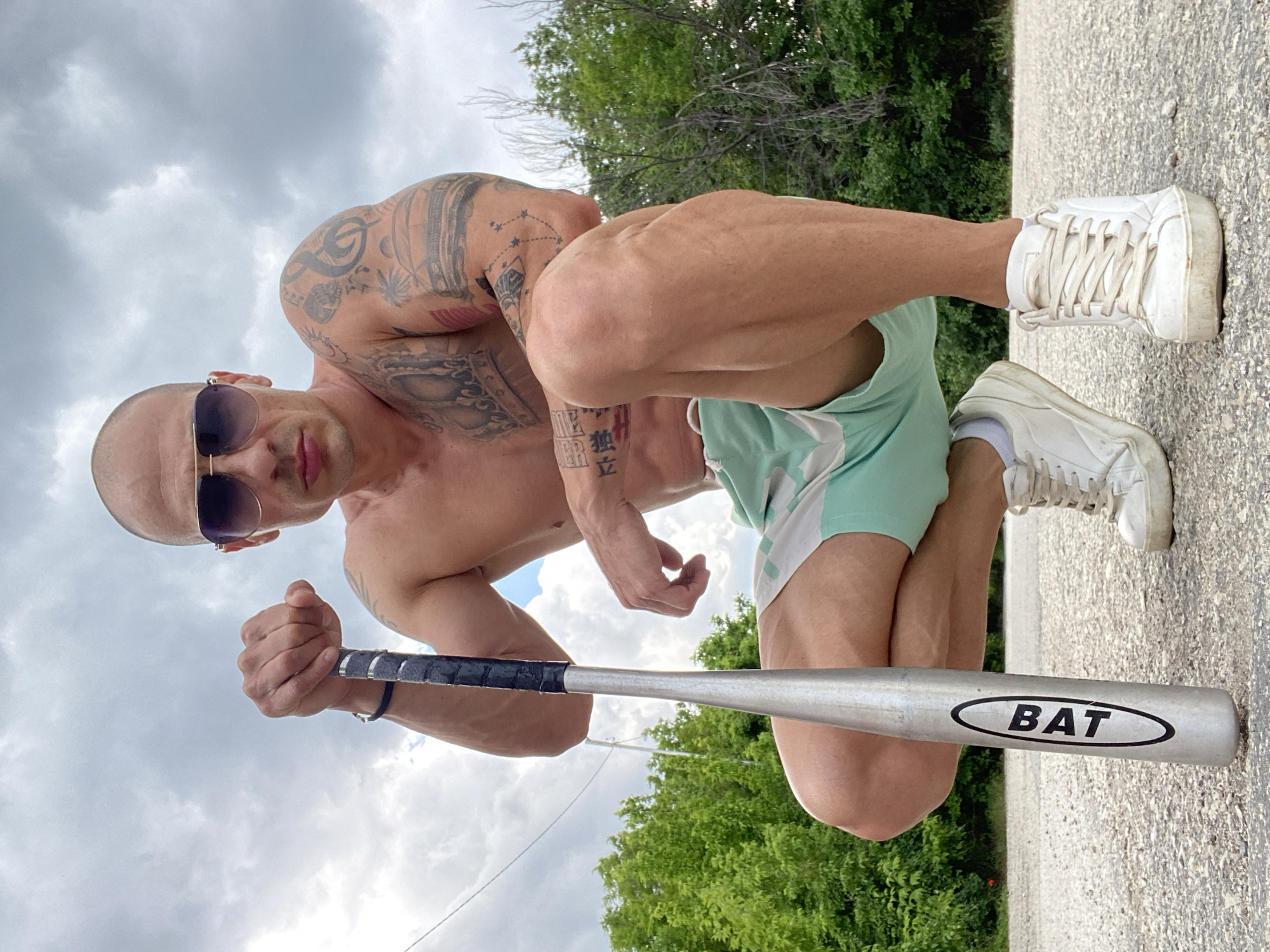 A shirtless, bald man with numerous tattoos on his arms and torso kneels on a paved surface. He wears sunglasses and light green shorts with white accents. He holds a silver baseball bat with a black grip. The word "BAT" is printed on the bat. He is wearing white sneakers. In the background is green foliage, possibly trees and bushes. The sky is overcast with a mix of clouds. It is daytime. The location and city are not discernible from the image.