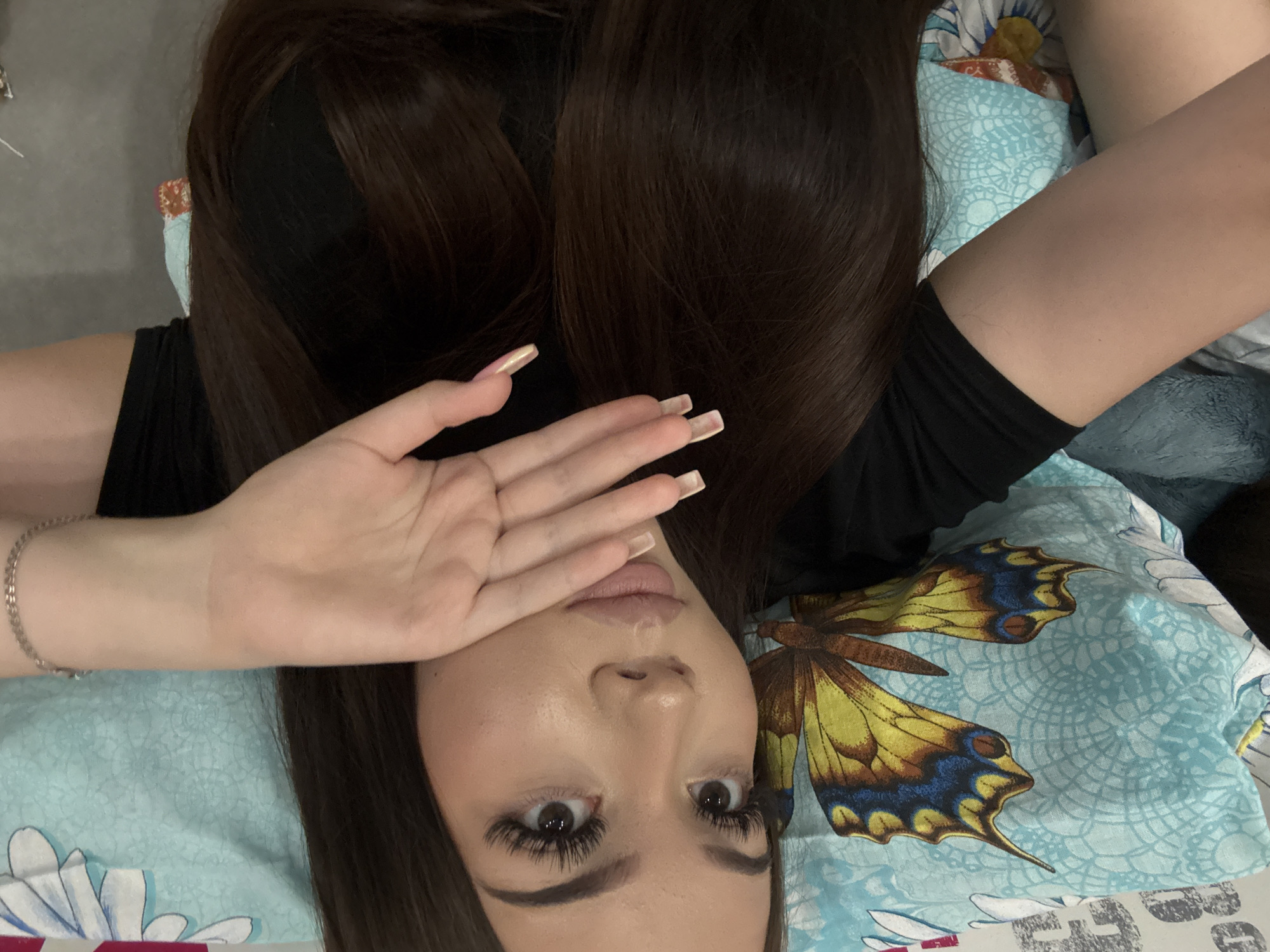 A young woman with long, dark brown hair and full makeup, including dramatic eyelashes, lies on a bed. She is wearing a black t-shirt. Her right hand is raised, covering her mouth and cheek, with long, light-colored fingernails. Her left arm is bent at the elbow, with a silver bracelet on her wrist. The background features a light blue pillow with a butterfly print and other bedding with floral patterns. The setting is indoors, likely a bedroom. The image was taken in Naberezhnye Chelny, Russia.