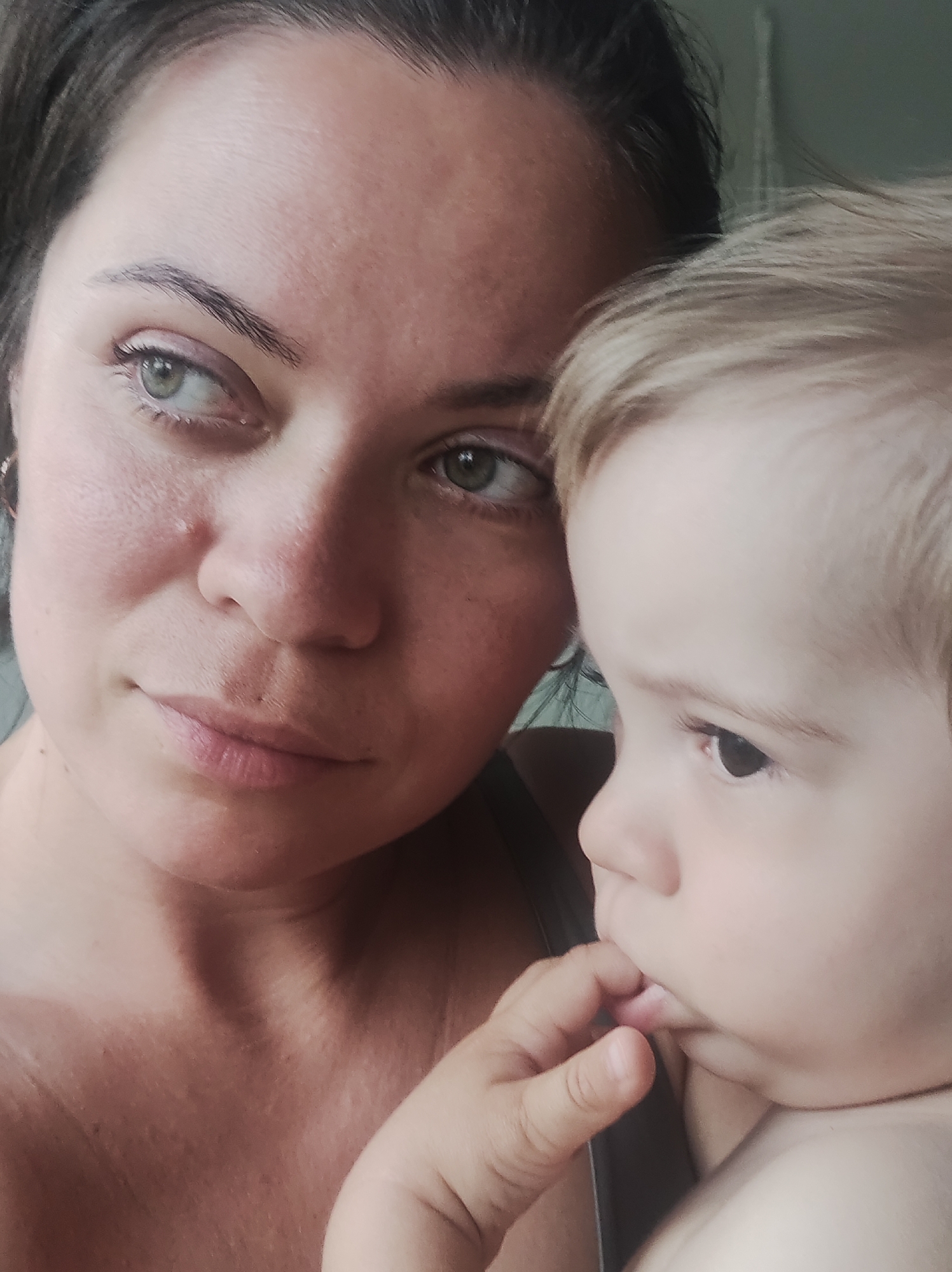 A close-up shot shows a woman and a baby, both looking towards the right side of the frame. The woman has dark hair pulled back, fair skin, and green eyes. The baby has light blond hair, fair skin, and is sucking on its fingers. The background is out of focus and appears to be an interior space. There are no indicators as to the location of the photograph, nor can the city of Chaikovsky, Russia be discerned from the image.