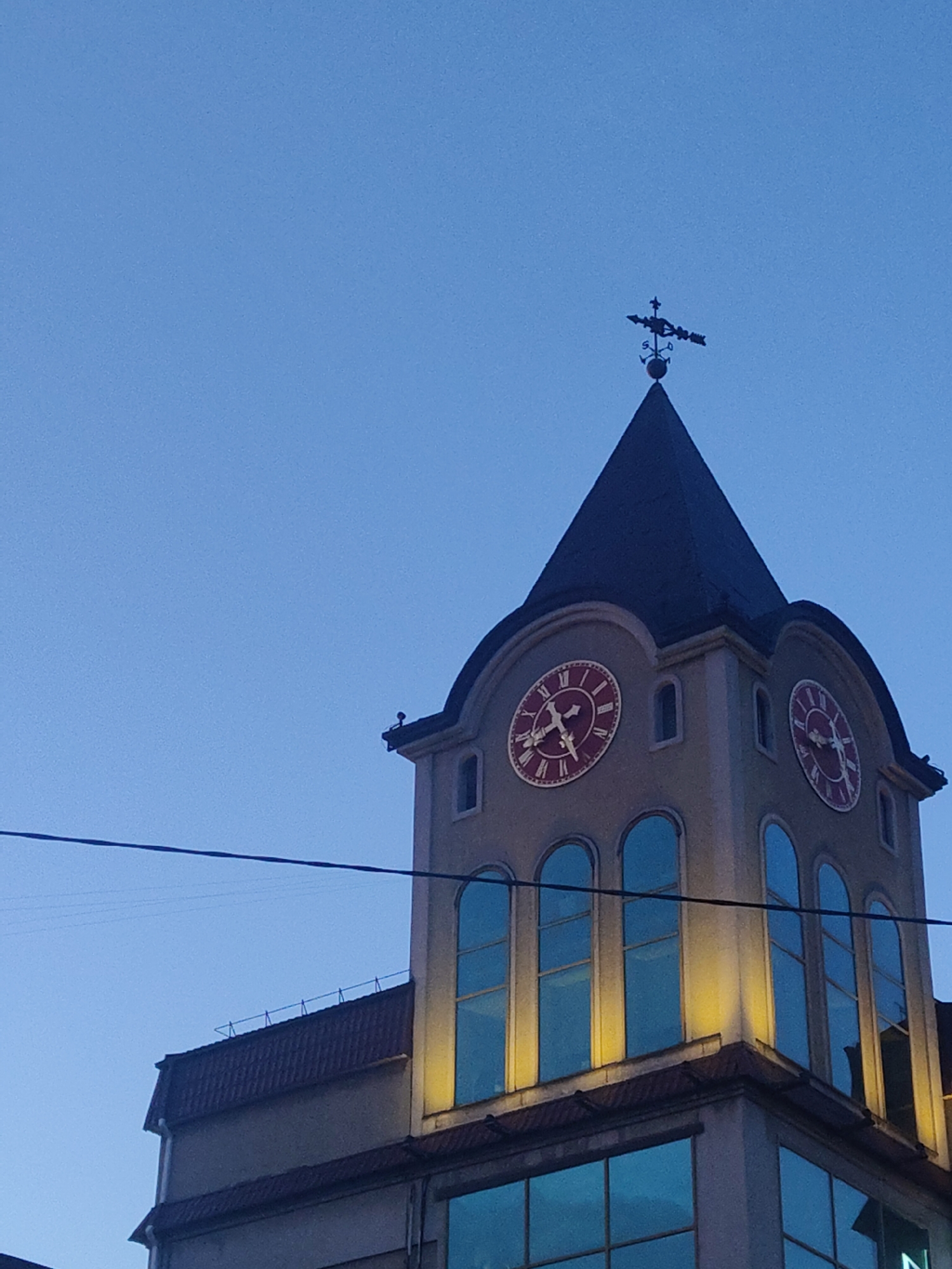 The image presents a striking view of a clock tower and a section of an adjoining building set against a clear, deep blue sky, suggesting dusk or early evening. The tower itself is a light beige color, featuring a dark, steeply pitched conical roof, which is topped with a distinct weathervane displaying cardinal directions (N, S, E, W) and an arrow.
Two prominent clock faces are visible on the tower, each recessed within an arched opening. Both clocks feature red Roman numerals, red minute and hour hands, and red details against a lighter background. The left clock indicates approximately 4:45, while the right one shows around 4:55.
Below the clock faces, the tower is adorned with multiple tall, arched windows that have a reflective blue tint, likely glass. Beneath these, warm, yellow artificial lights illuminate the building's facade. A darker, possibly tiled, roof section of an adjacent structure is visible to the left of the tower. The lower part of the image reveals large rectangular windows with similar blue reflections on the building's ground floor. A dark power line stretches horizontally across the mid-section of the frame. No people are visible in the scene. The architectural style appears to have classical or European influences.