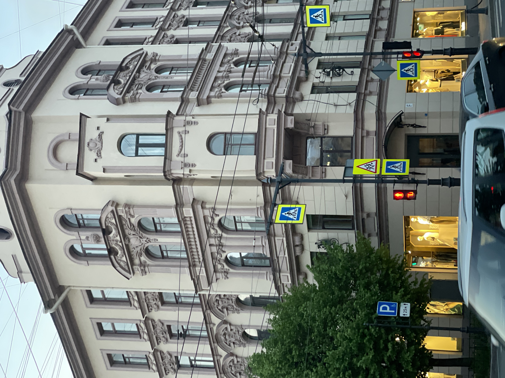 The image shows a street scene in Saint Petersburg, Russia. An ornate building facade with arched windows and decorative moldings dominates the left side of the frame. Street signs, including pedestrian crossing signs and a parking sign, are visible along with traffic lights showing red. A tree is present in the lower-right corner. Cars partially obstruct the view, suggesting an active street. Shop windows are also visible in the background.
