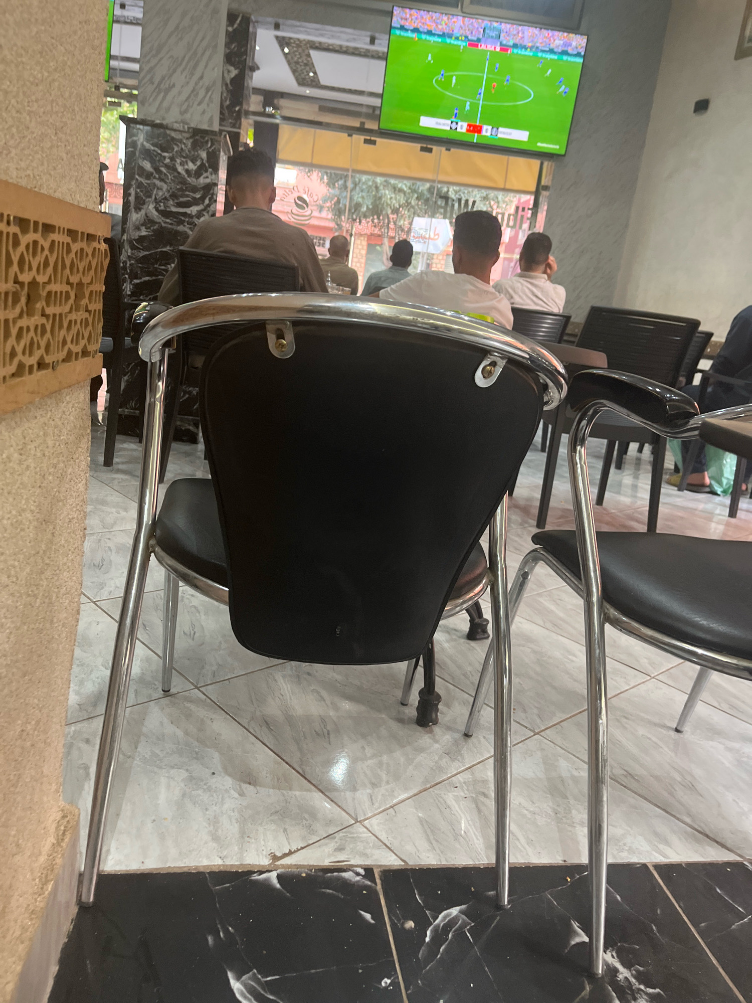 The image depicts the interior of a cafe or restaurant in Azilal, Morocco, where patrons are watching a soccer match on a large television screen. In the foreground, two chairs with black seats and chrome legs are visible. The chair on the left is closer to the camera, with its backrest facing away. The chair on the right is partially obscured.
Several people are seated in the cafe, their backs mostly to the camera, as they focus on the game. One individual in the middle background wears a white t-shirt. Further back, other patrons are seated at tables, also engaged with the television. The cafe features tiled flooring and marble-patterned pillars. A decorative wooden lattice is visible on the left side of the image. The overall atmosphere suggests a casual gathering of people enjoying a sporting event.