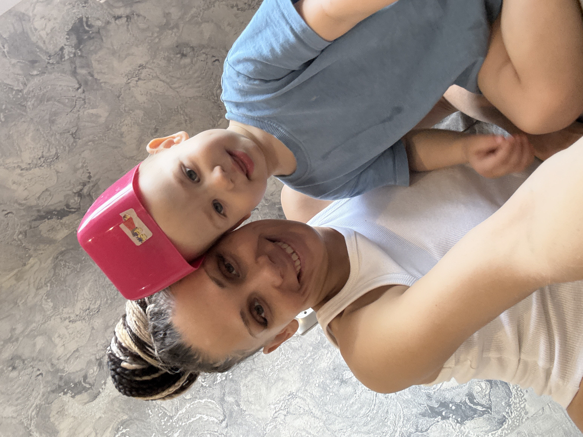 A woman and a child are in a selfie-style photograph. The woman is wearing a white tank top and has her hair up in braids. The child is wearing a blue shirt and has a pink plastic object on his head. They are both smiling at the camera. The background appears to be a textured wall. The location is not discernible from the image alone. I cannot determine the city of Yaroslavl', Russia as a location.
