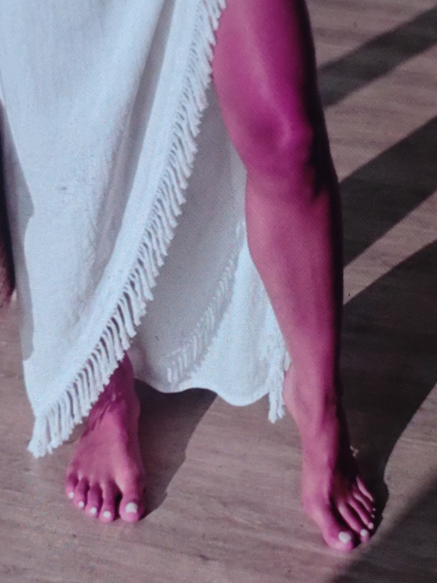 In this close-up, low-angle shot, a person's legs and feet are visible, bathed in a vibrant pink light. The left leg is partially obscured by a white garment with fringe detailing, while the right leg is fully visible, its skin appearing a deep magenta. The person is barefoot, standing on a textured surface that appears to be wood or a similar material. Shadows cast by parallel lines create a striped pattern across the ground, suggesting the presence of blinds or a slatted structure. The overall lighting is dramatic, highlighting the contours of the leg and feet.