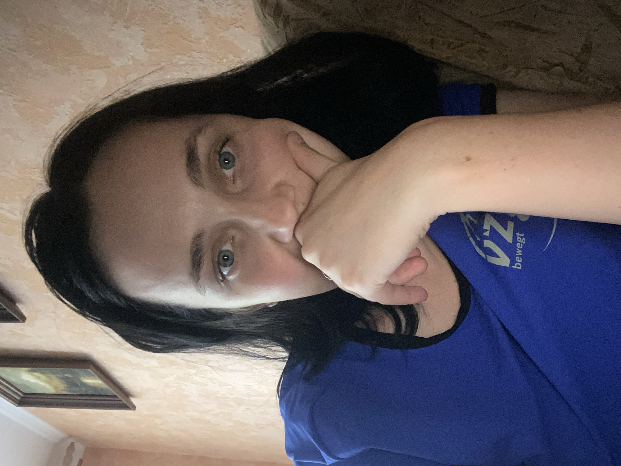 A woman is taking a selfie, with her face partially obscured by her hand. She has dark hair and blue eyes and is wearing a blue t-shirt with a logo on it. The background appears to be a wall with a textured finish and a framed picture. The lighting is dim, suggesting an indoor setting. There is no identifiable information about the location within a city.