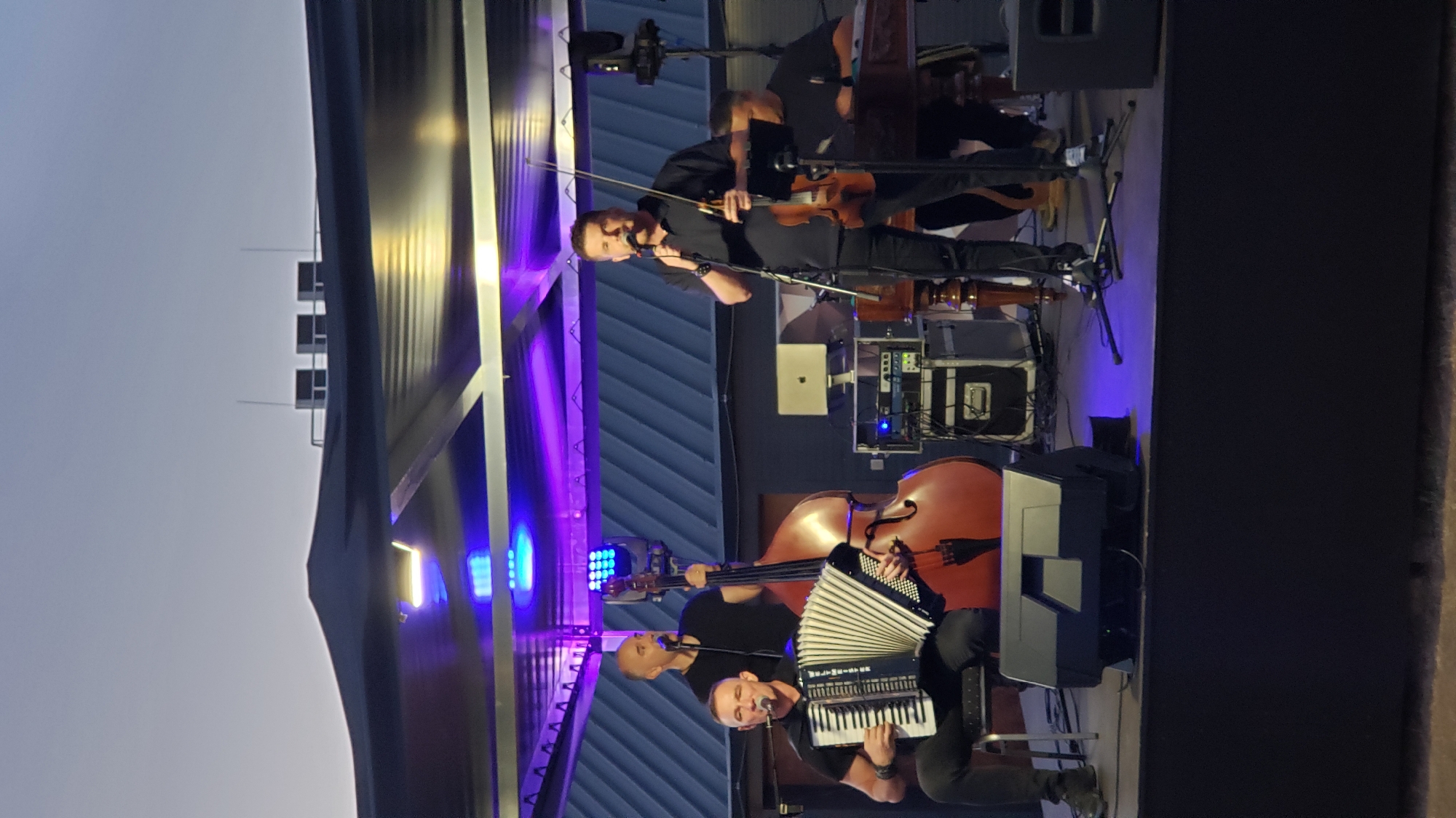 A band performs on an outdoor stage under a canopy at dusk. In the foreground, a man sits playing a white accordion, his arms extended. To his right, a double bass stands vertically, and behind it, a man stands holding a microphone, singing. In the center of the stage, another man stands holding a violin and bow, also singing into a microphone. To the right, a man is seated, appearing to play a keyboard or similar instrument. The stage is lit with purple and blue lights. In the background, a metal structure with ventilation units is visible against a twilight sky. The overall setting suggests a performance venue in the city of Prešov, Slovakia, possibly an outdoor concert or festival.