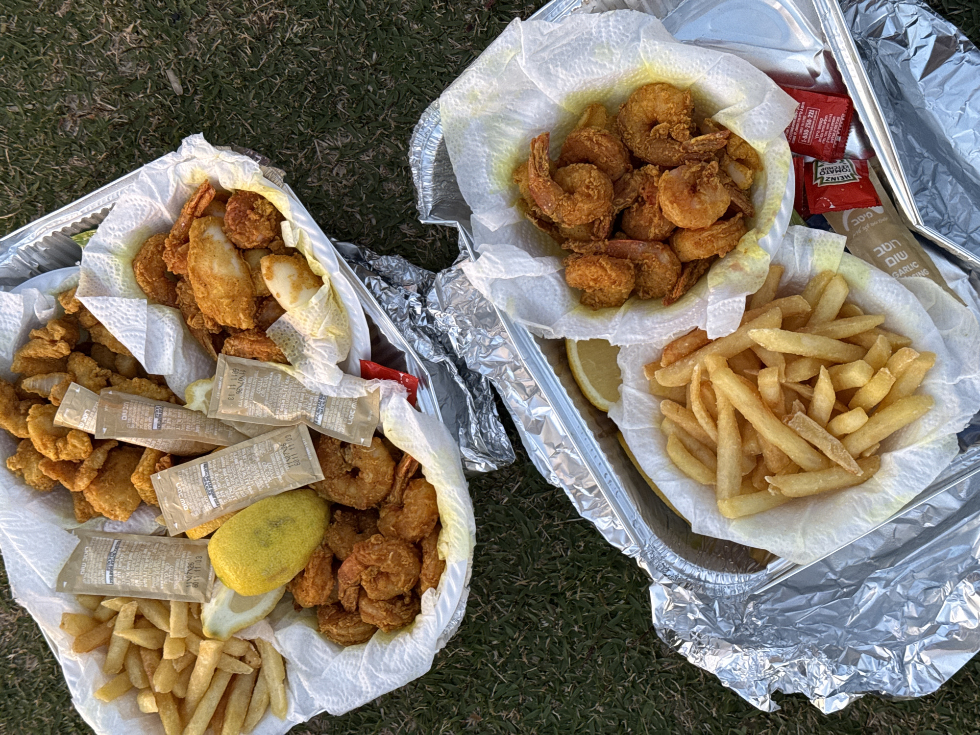The image shows two aluminum trays filled with fried food on what appears to be grass. The tray on the left contains fried fish, shrimp, and french fries, with several packets of garlic sauce and a lemon wedge on top. The tray on the right contains shrimp, french fries, a lemon wedge, and packets of garlic sauce and ketchup. The labels on the sauce packets are in Hebrew. The food appears to be a takeout meal. Considering the Hebrew writing and the style of food, it is likely that the scene is set in Tel-Aviv, Israel.