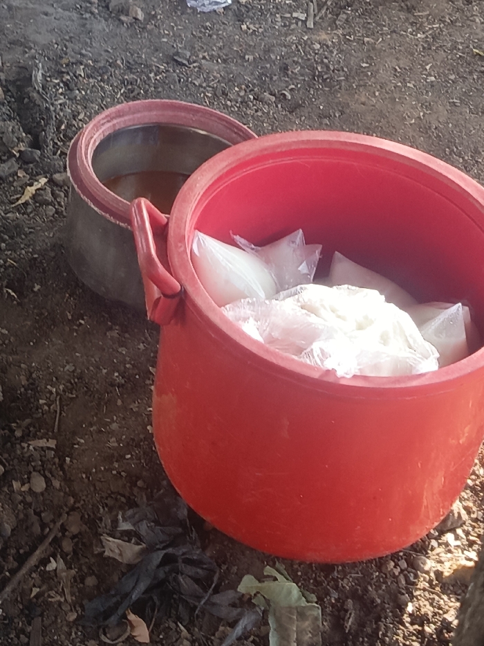 Two round containers sit on dirt and debris. The container in the foreground is red and filled with white, plastic-wrapped items. Behind it is a larger, grey container filled with a brownish liquid. The ground surrounding the containers is covered in bits of sticks, leaves, and discarded plastic bags. The containers appear to be outdoors, possibly in a rural or underdeveloped area.