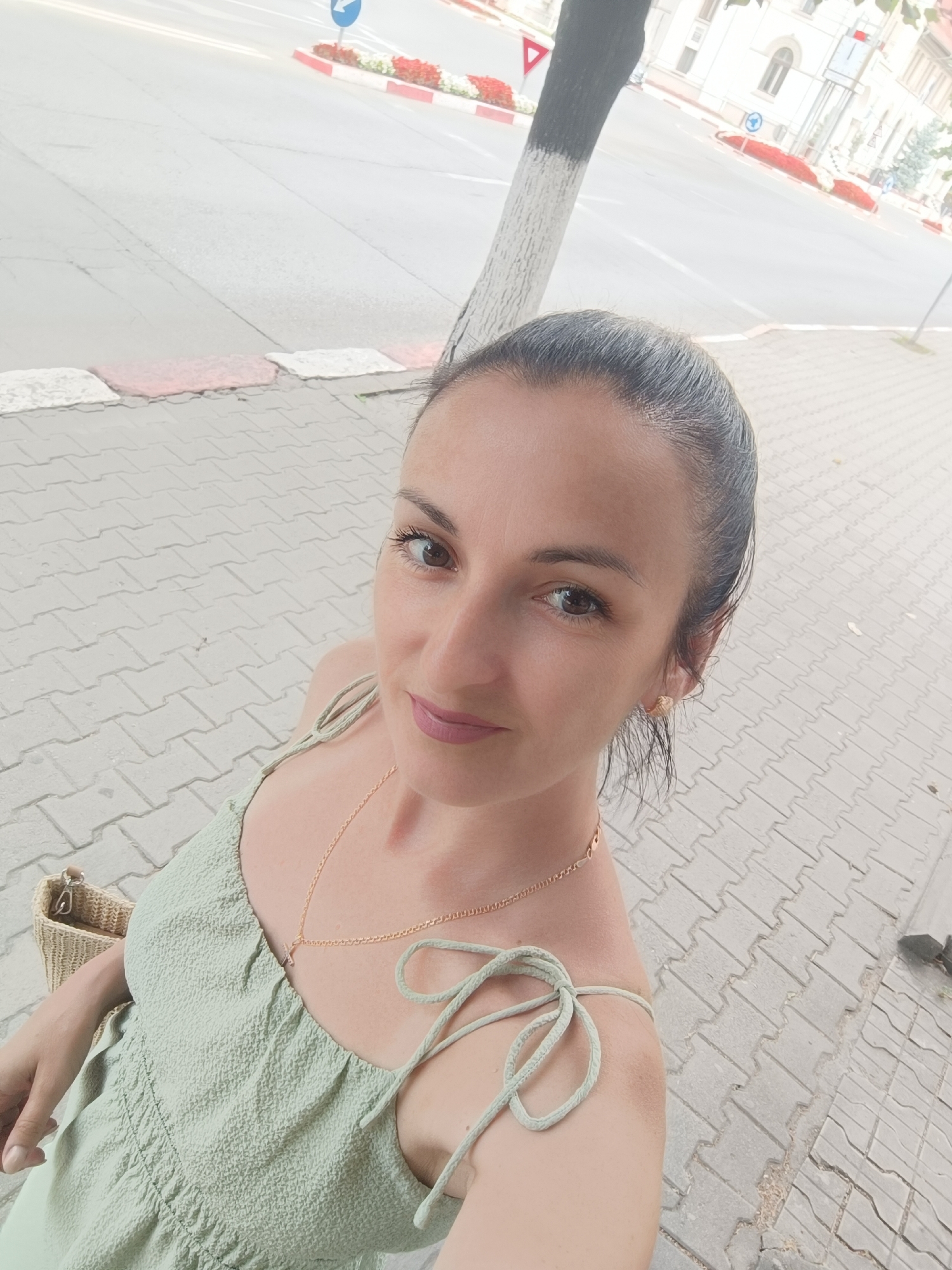 A woman with dark hair pulled back in a ponytail takes a selfie outdoors. She is wearing a light green dress with tie straps and a gold necklace. Her right hand is resting on her chest. To her left, a straw bag is visible. She is standing on a paved walkway with a street and buildings in the background. The environment suggests an urban setting, possibly in Târgu Jiu, Romania, given the presence of a tree with black and white painted markings on its trunk, a common practice for marking trees near roads in some European countries. The walkway is made of interlocking grey pavers. A street with red and white painted curbs runs horizontally in the upper portion of the image.