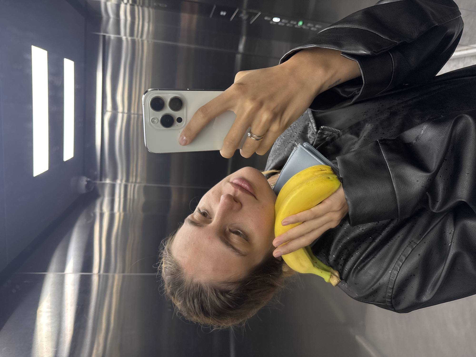 A woman is standing in an elevator, taking a selfie with a smartphone. She is holding a bunch of bananas against her chest and wearing a black leather jacket. The elevator has metal walls with three vertical light fixtures. The location is Unknown, Unknown.