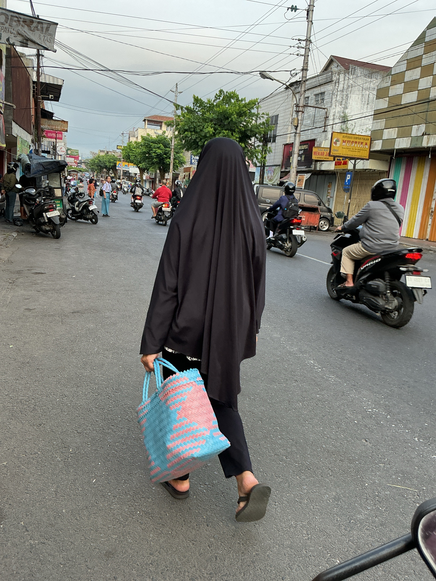 The image captures a street scene in Magelang, Indonesia, with a focus on a person from behind wearing a black hijab and carrying a blue and pink woven bag. The individual is walking on a paved road, which appears to be a commercial area, with several motorcycles parked and moving around. Shops and buildings line the street, some with visible signage. The sky is overcast. Other people on motorcycles are also present in the scene.