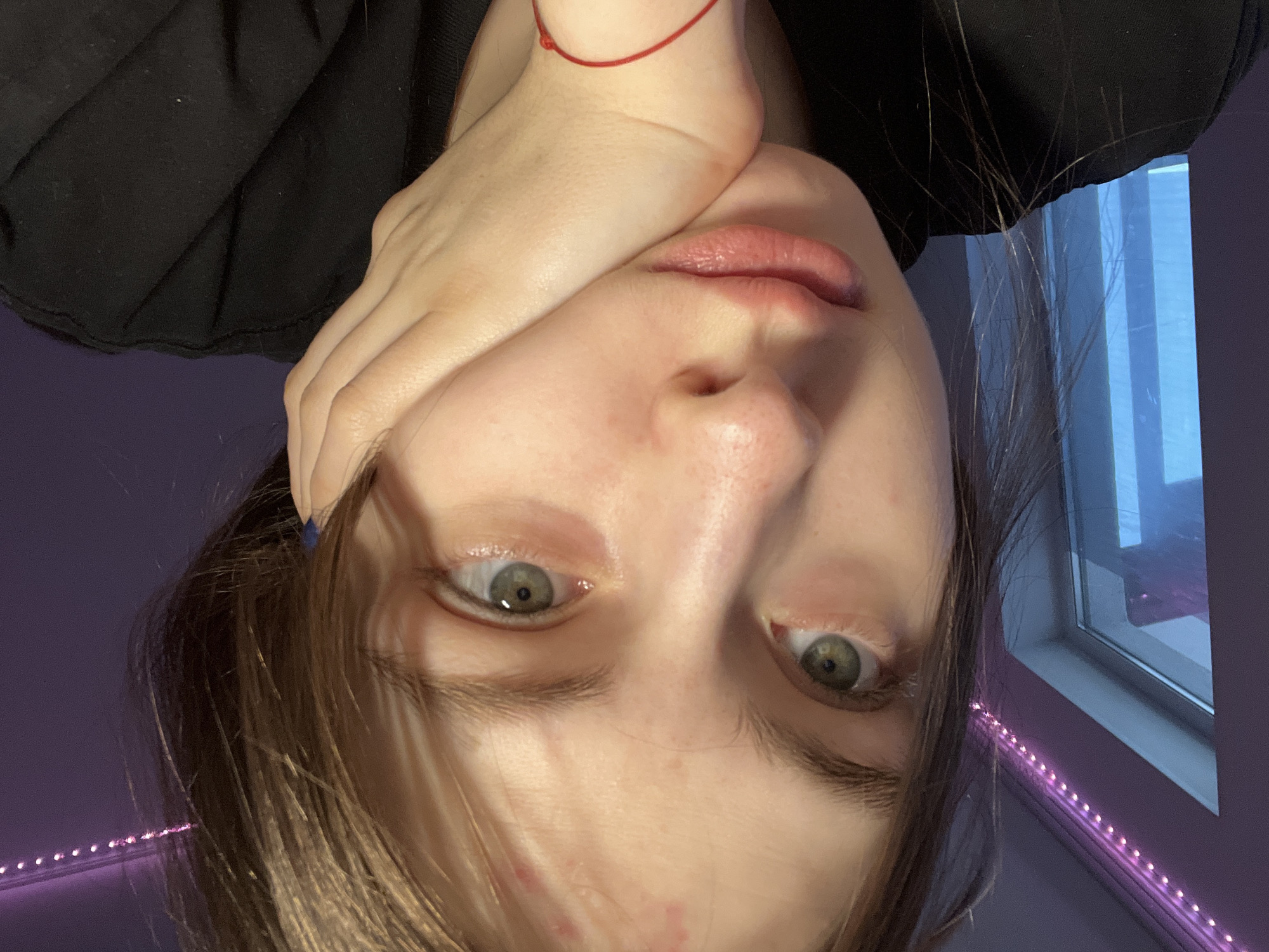 A portrait photograph, rotated 180 degrees, depicts a fair-skinned individual with green eyes, light brown hair, and a hand partially covering the face. A red string bracelet is visible on the wrist. The person is wearing a black top. The background includes a window frame and purple wall illuminated by a string of pink LED lights. There is no indication the location of this image is Saint Petersburg, Russia.