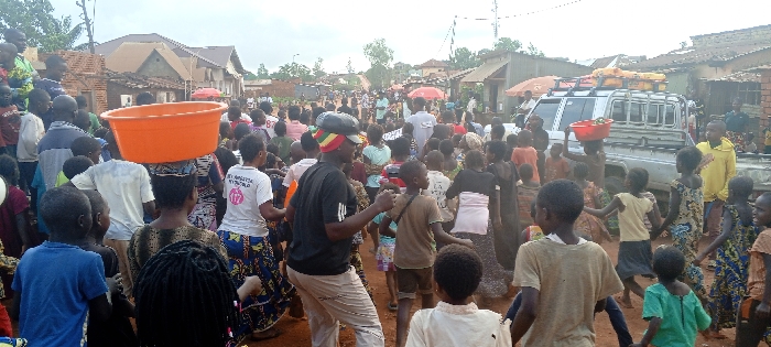 The scene depicts a dense crowd of approximately one hundred individuals, primarily children and adults of African descent, occupying an unpaved, reddish-brown dirt street or open area within the city of Fizi, Democratic Republic of the Congo.
In the foreground and midground, numerous children are present, many appearing to observe or move within the crowd. Adults are interspersed throughout. A woman in the left midground carries a large, empty, orange plastic basin balanced on her head. Towards the center, a male figure wearing a black t-shirt with white stripes on the sleeves, light-colored pants, and a black cap with red, yellow, and green stripes is visible, appearing to be in motion with his arms slightly raised. Another person in the right midground carries a smaller red basin containing green items, also balanced on their head.
In the right midground, a light-colored pickup truck with a utility bed is parked or moving slowly. Its roof rack is laden with several yellow jerry cans or containers and other indeterminate items.
The background features numerous single-story and two-story structures with brick walls and various roof types, indicating a built-up urban or semi-urban environment. A red market umbrella is partially visible behind the truck. The sky is overcast and grey. Utility poles and overhead lines are discernible in the distant background.