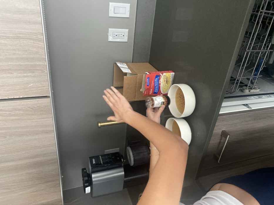A person's arms add spice to soup in two white bowls, next to a box of Lipton Extra Noodle soup. Adjacent to the bowls is an open dishwasher. To the left of the bowls, a brown cardboard box is visible. A Ninja blender and gold handle are below these items. Above are an electrical outlet and a light switch. A wood-grain cabinet is to the left, and a gray cabinet encompasses the background.