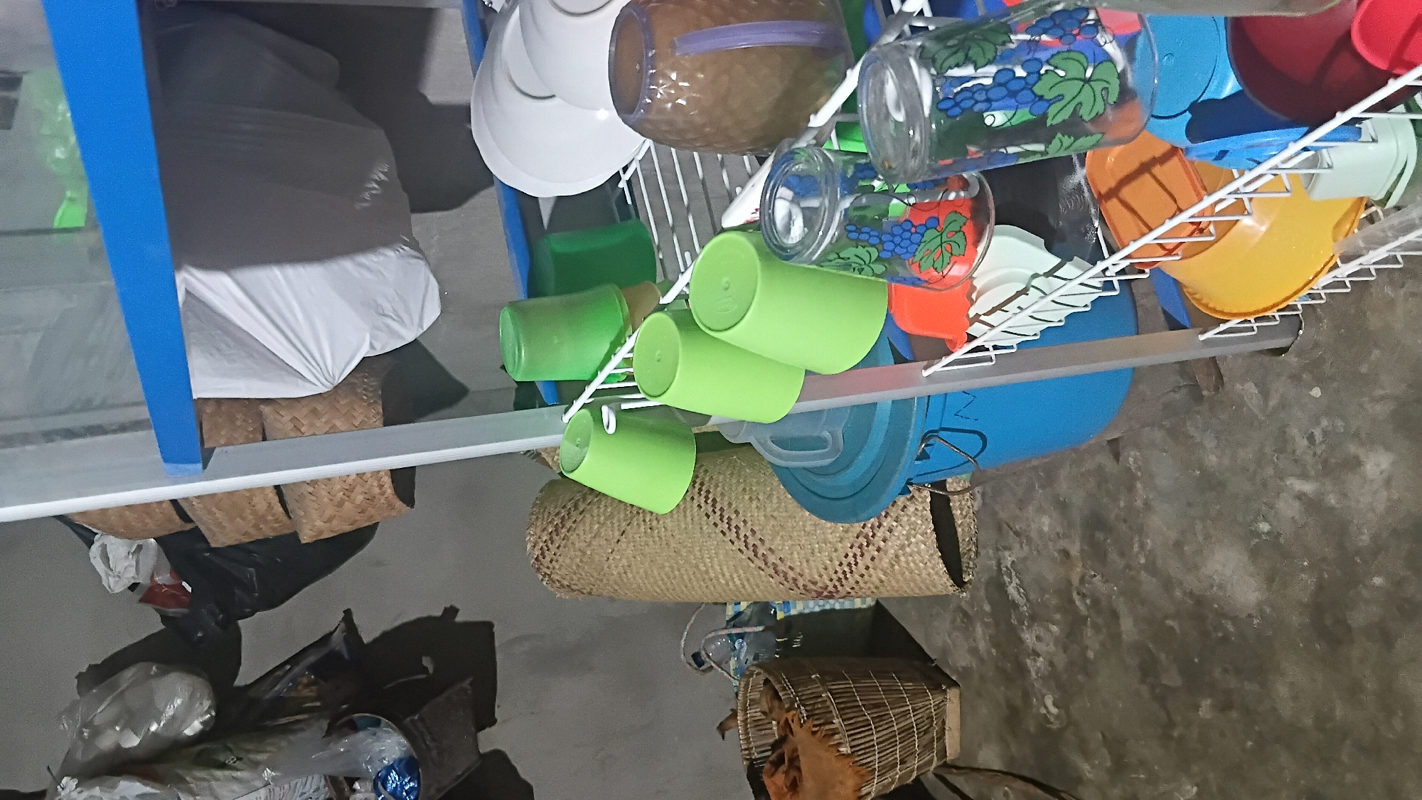 A domestic scene is depicted in a cluttered room with concrete floors and walls. A white shelving unit with blue trim holds various kitchen items. On the shelves are stacks of white plates, a brown patterned pitcher, several green plastic cups, and clear glass jars decorated with grapes. Below the shelves, a large blue plastic bucket and a yellow bowl are visible. To the left of the shelving unit, a woven mat stands upright, leaning against the wall. A wicker basket filled with fabric and a rolled-up mat is also present. Bags and other items hang on the wall in the background. The scene appears to be an indoor storage area or kitchen in a residential setting, possibly within the city of Prabumulih, Indonesia, given the context.