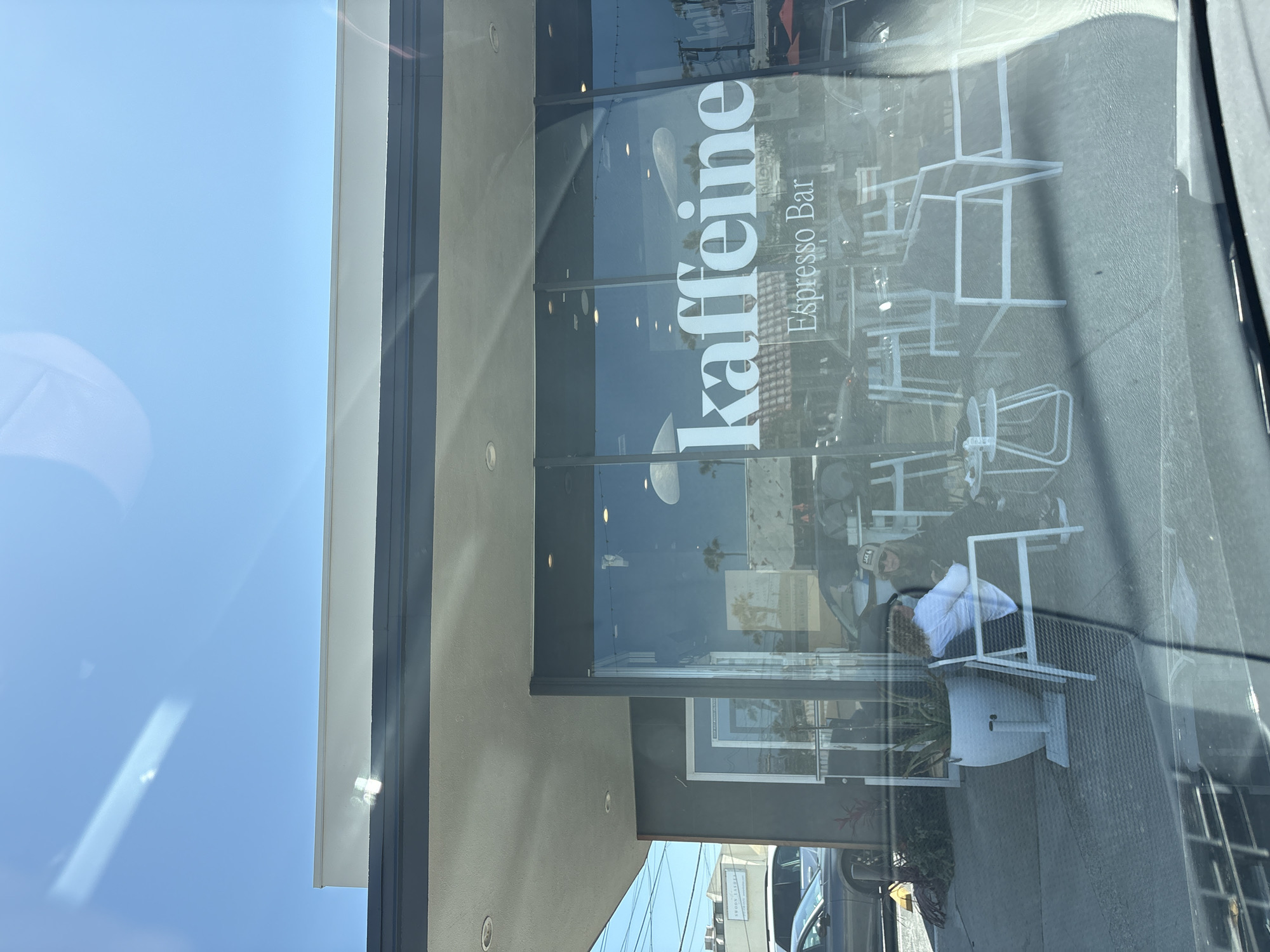 Here's a forensic description of the image:
**Location:** Redondo Beach, California, United States of America.
**Subject:** A storefront for "Kaffeine Espresso Bar".
**Exterior:**
* The building has a modern architectural style with a flat roof, grey wall finish.
* A glass storefront is prominently displayed with the business name "Kaffeine" vertically oriented in white letters, along with "Espresso Bar" printed beside the business name.
* Through the glass, white outdoor seating can be seen, which includes tables and chairs.
* Reflections on the glass indicate the presence of traffic, other businesses, and some greenery across the street.
**Interior:**
* The interior is visible through the glass, suggesting a cafe-style setup with chairs and tables.
* Patrons are seated and appear to be engaged in conversation.
**People:**
* Individuals can be seen seated at tables through the glass window, inside the café.
**Objects:**
* Cars are reflected in the glass.
* The business name "Kaffeine" in large, white letters.
* White cafe-style chairs and tables.
* Exterior plants.
**Environment:**
* The environment is urban, suggesting a business district or street with pedestrian activity.
* The daylight indicates a sunny day.
**Inferences:**
* The storefront belongs to a coffee shop named "Kaffeine Espresso Bar".
* It is an active business with patrons inside.
* The business appears to have outdoor seating.