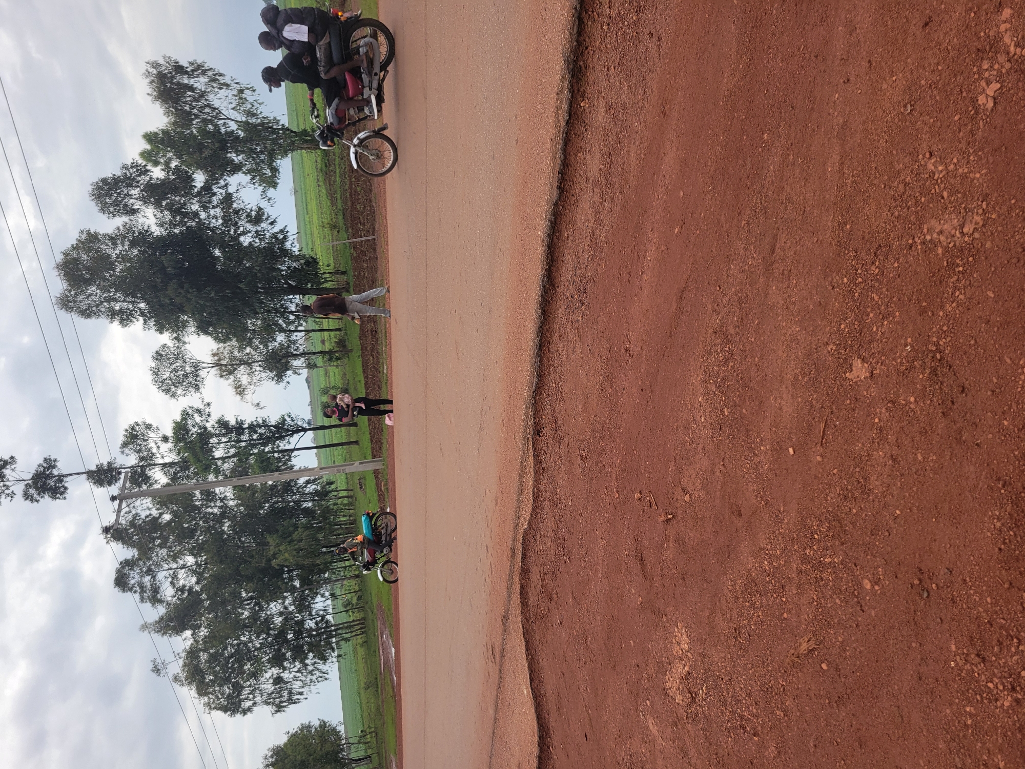 A horizontal, eye-level shot shows a rural road with a dirt shoulder and a paved surface. Several tall, slender trees with sparse foliage line the background, with a field of green crops visible beyond them. A power line stretches across the top of the frame, supported by a concrete pole.
In the mid-ground, several people and a motorcycle are present. A woman in black pants and a black top holds a baby. A man in a brown shirt and light-colored pants walks away from the camera. A motorcycle with three occupants is riding on the paved road. The driver wears a dark helmet and a light-colored vest over a dark shirt. The two passengers behind him wear dark clothing. A second motorcycle, partially obscured by the first, is parked on the side of the road, with its rider wearing a high-visibility vest.
The foreground consists of a red dirt surface with scattered gravel and pebbles. The road itself appears to be made of reddish asphalt. The sky is overcast with diffused light.