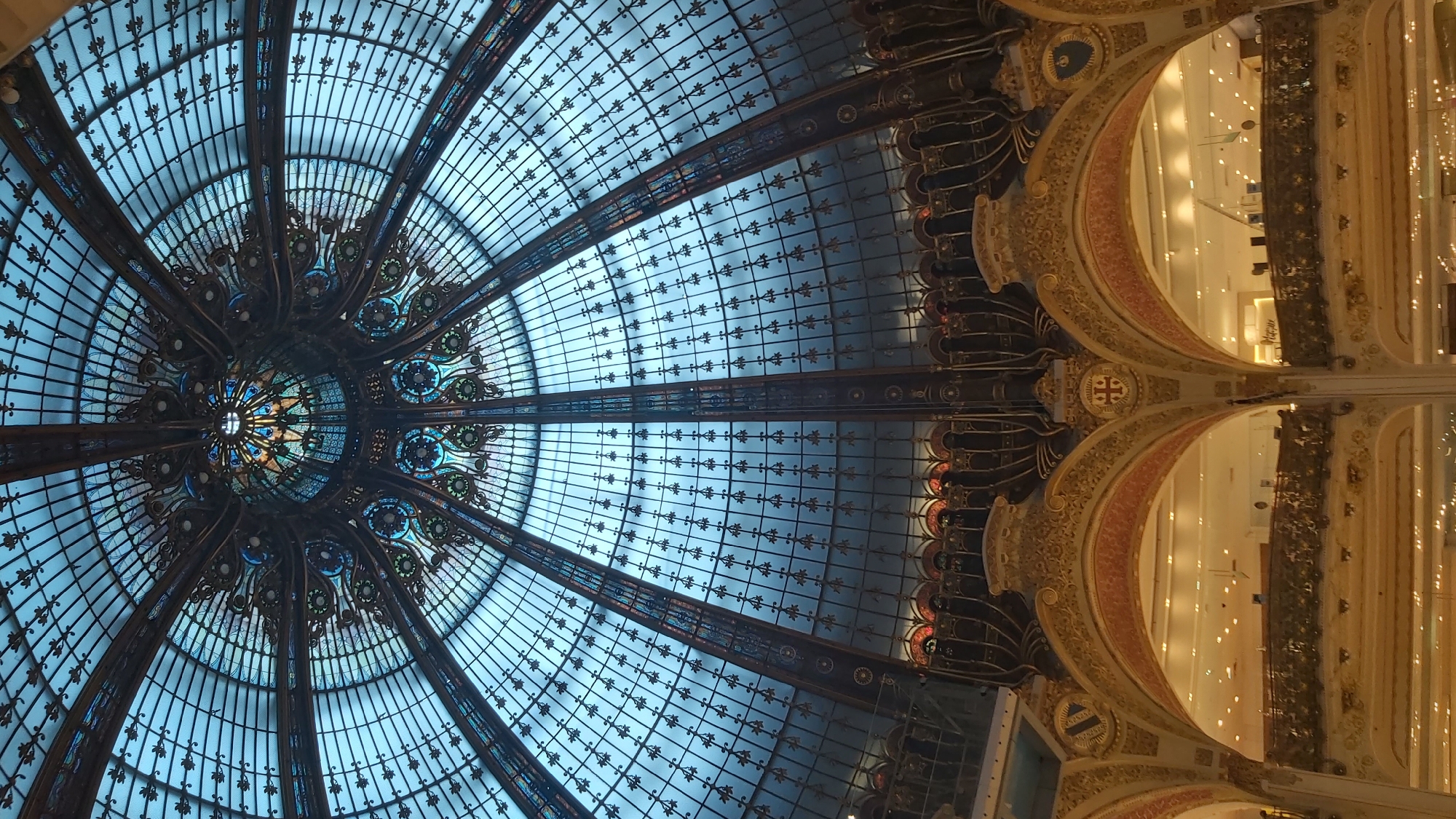 This image depicts the ornate Art Nouveau dome of the Galeries Lafayette Haussmann department store in Paris, France. The dome is constructed of intricate stained glass and wrought iron, with geometric patterns radiating outwards from a central, colorful medallion. Below the dome, elaborate golden arches and balconies, adorned with decorative carvings and crests, frame views of the brightly lit interior of the department store, where shoppers can be seen on various levels. The overall impression is one of grandeur and architectural splendor.
