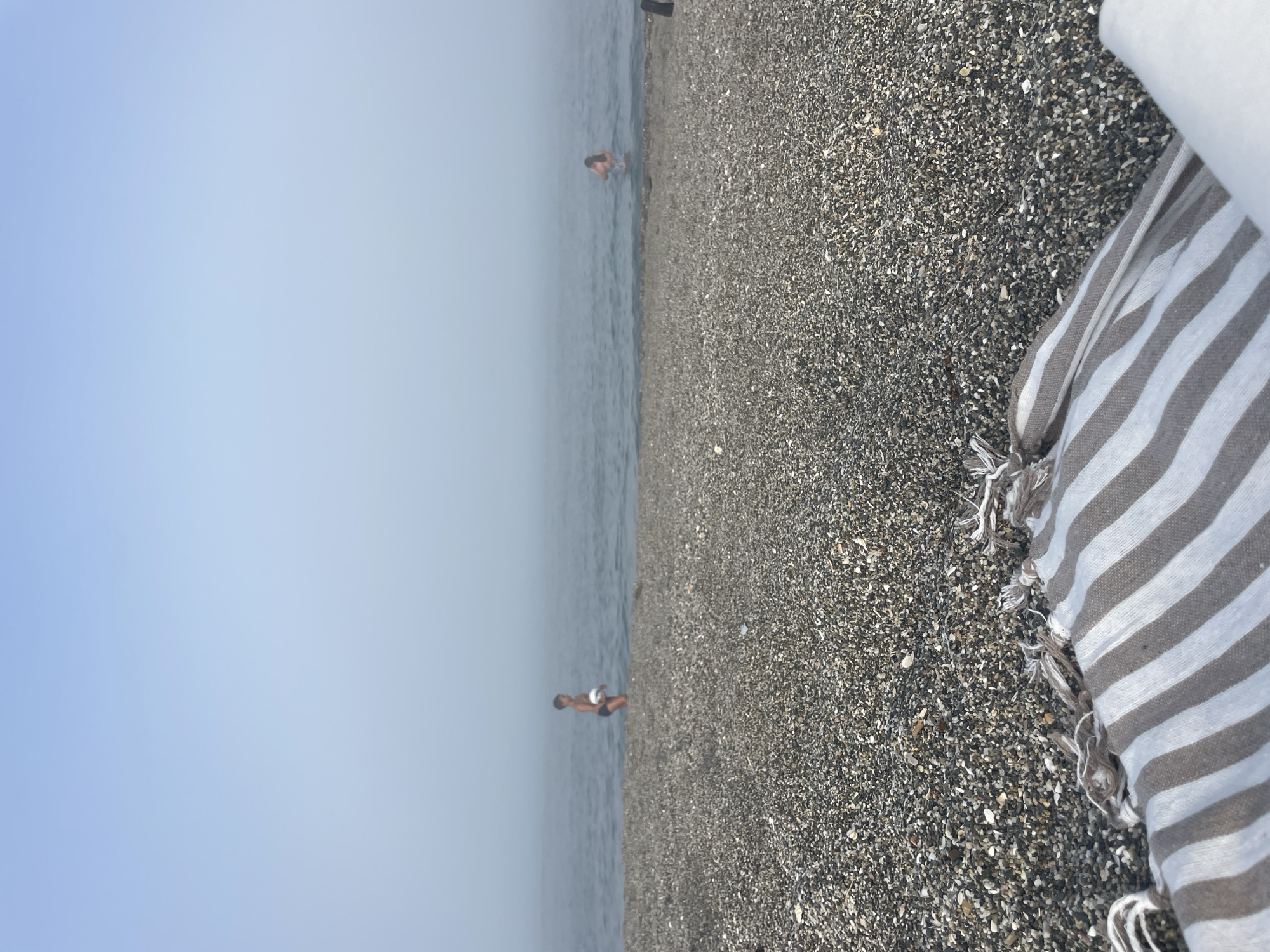 The image depicts a serene beach scene with a focus on a person standing in shallow water and a striped towel draped on the pebbled shore. The horizon is barely visible, and the sky is a hazy, pale blue, suggesting either early morning or late afternoon. The water is calm and a muted grey-blue, with small ripples on its surface. The beach is covered in dark grey pebbles and small stones.
In the distance, a person is wading in the sea, their body partially submerged in the water. They appear to be a young individual wearing dark swim trunks. The towel in the foreground is made of a light fabric with alternating white and grey stripes. Its fringe is visible, adding a tactile element to the scene. The composition is such that the beach and towel occupy the right two-thirds of the frame, while the sea and sky fill the left third, creating a sense of depth and vastness.
There are no discernible signs of other activity or specific events, beyond the simple act of being at the beach. The overall mood is peaceful and tranquil. The light is soft, and there is no strong indication of harsh sunlight. The location cues are minimal, but the presence of a beach and the clear, calm sea are evident. No text is visible in the image. The scene evokes a sense of quiet relaxation and enjoyment of a coastal environment.