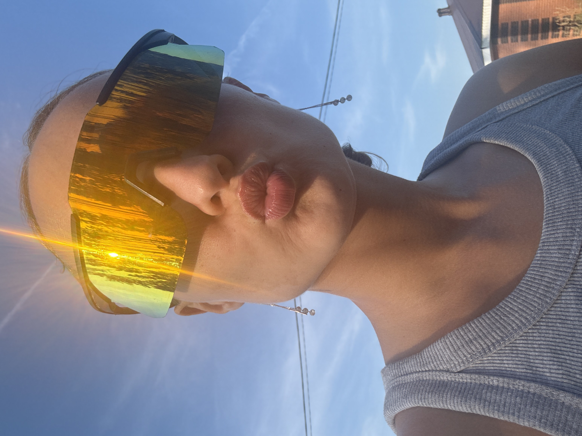 A woman, with her lips pursed, poses for a selfie outdoors against a bright blue sky with wispy clouds. She wears large, reflective, yellow-tinted sunglasses and a grey ribbed tank top. Long, delicate earrings with small dangling charms hang from her ears. The sun glints brightly off her sunglasses, creating a halo effect. In the background, a portion of a brick building with a dark roof is visible, suggesting an urban or suburban environment. The image captures a moment of leisure on a sunny day, possibly in the city of Podshivalovo, Russia, given the context.