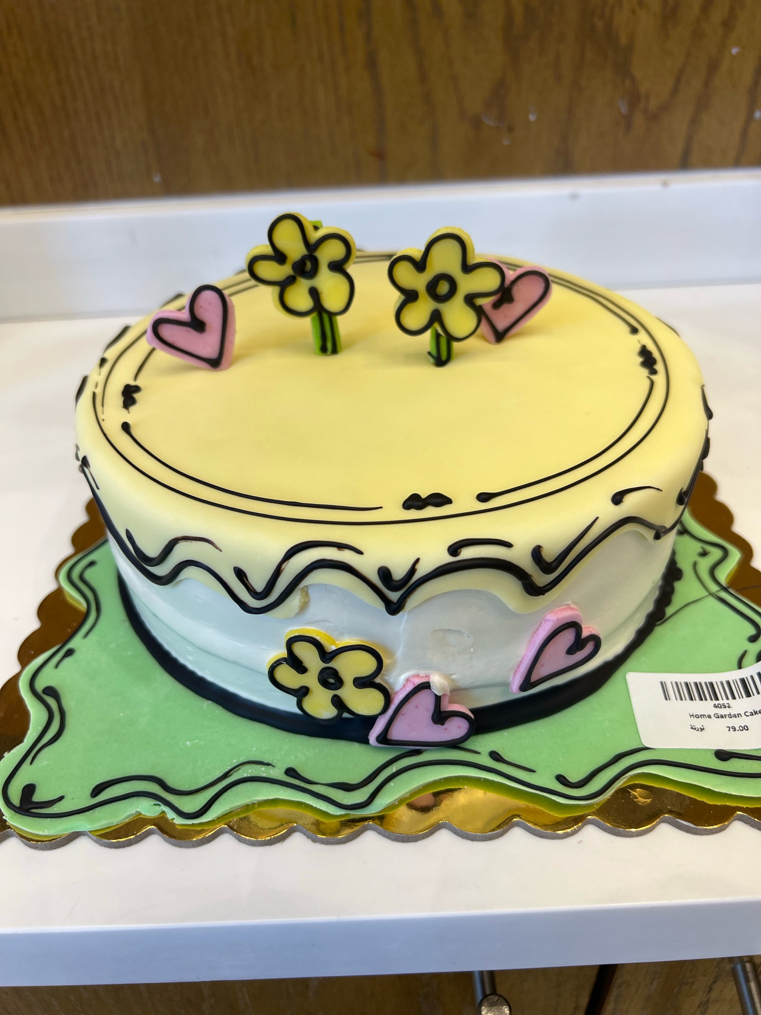 A round, two-tiered cake is presented on a decorative green and gold base. The top tier is covered in yellow frosting and adorned with black line drawings, two yellow flowers with green stems, and three pink hearts. The bottom tier is frosted in white and decorated with a black wavy pattern and several pink hearts and yellow flowers. A white label with a barcode is affixed to the side of the cake base. The cake sits on a white surface, with a wooden background visible behind it. This image was captured in Al Khobar, Saudi Arabia.