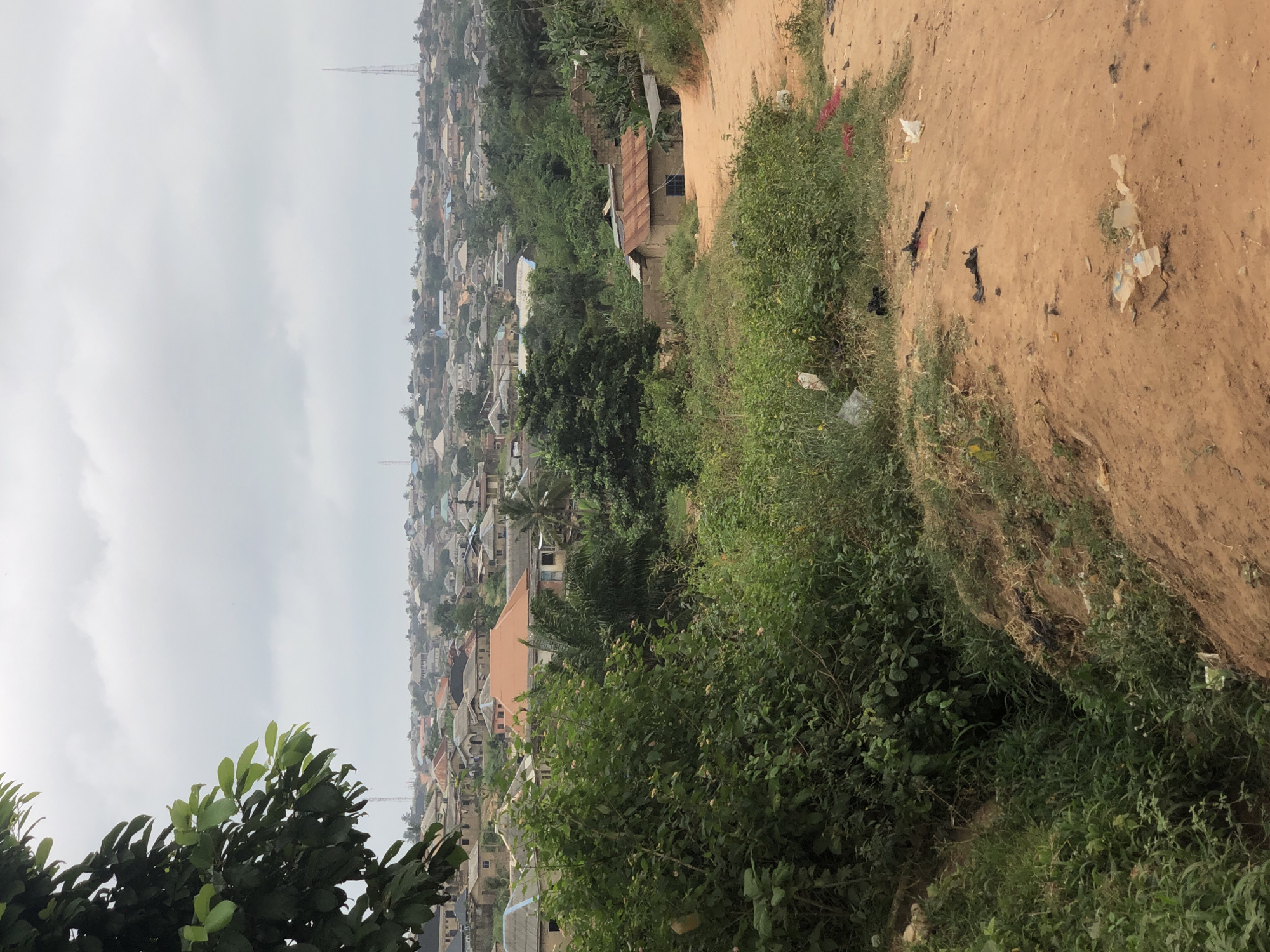 The image captures a panoramic view of a densely populated urban settlement in Ijoko, Nigeria, under a cloudy sky. The foreground is dominated by lush green vegetation and a reddish-brown dirt path, with some scattered litter. A steep embankment of the path, covered in grass and foliage, is visible on the right. In the midground, several modest houses with corrugated iron roofs and some with concrete roofs are nestled among trees and greenery. The architecture suggests a mix of temporary and more permanent structures. In the background, a sprawling cityscape extends across rolling hills. Numerous buildings, predominantly single-story, are visible, interspersed with more vegetation. Two tall telecommunication towers rise above the horizon, indicating infrastructure development. The overall impression is one of a developing urban area with a significant population living in close proximity, surrounded by natural elements.