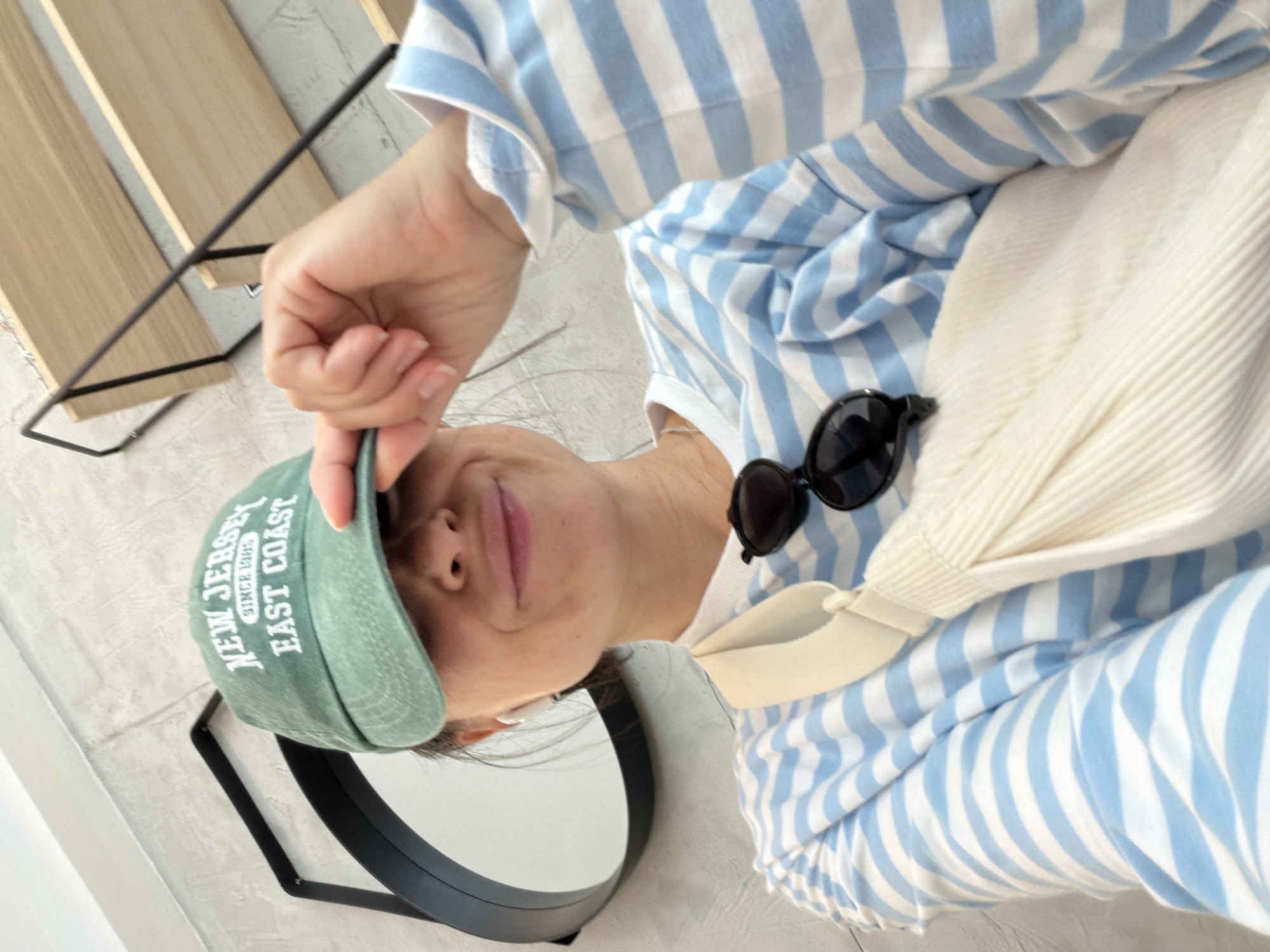 A woman in a blue-and-white striped top and green baseball cap is posing for a photograph. She is holding the brim of her cap. Sunglasses are clipped to her shirt, and she has a light-colored bag slung over her shoulder. A black-framed mirror is visible to the left of her face. A wooden shelf with black metal supports is in the background. The location of Budva, Montenegro is not discernible from the image.