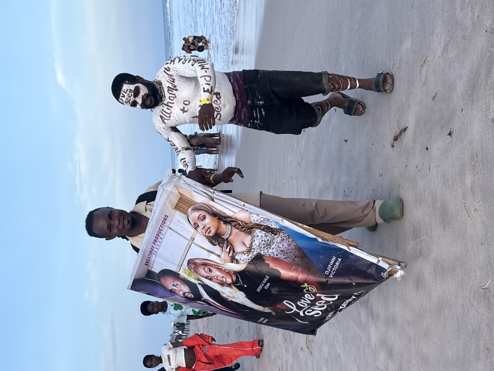 The image shows a promotional scene on a beach, likely in Mopo Onijebu, Nigeria, given the style of attire and promotional materials. In the foreground, two men are featured prominently. One man is wearing what appears to be body paint with writing on it. He has white paint covering his face and torso, with black writing over it, including words like "Allihamdul," "to," "Eid-Mu," and "Seed". He wears dark pants and sandals.
The second man holds a large, damaged promotional poster for a movie titled "Love Seed," featuring images of two women and a man with their names listed as Jessica Ikhile Edan, Ojifinni Victoria and "TOWING BASSRY". The poster reads "BREATHWAY PRODUCTIONS PRESENT". He wears khaki-colored pants and light green footwear.
The background reveals a beach setting with an ocean and sky. A group of people can be seen partially visible behind the men in the foreground. There are other people standing on the beach.