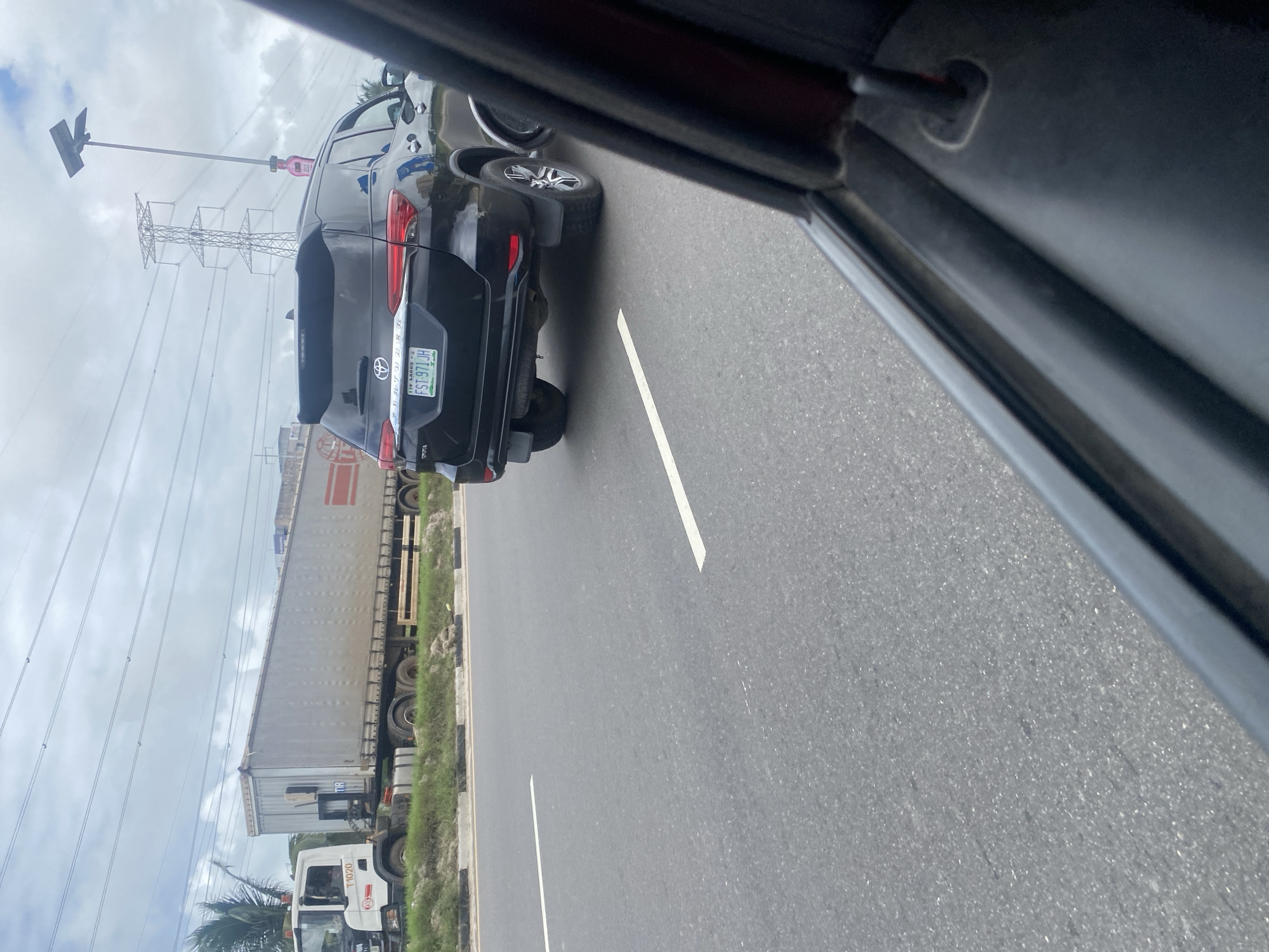 The image shows a black Toyota Fortuner driving on an asphalt road in Ilado, Nigeria. The vehicle is in motion, indicated by the slight blur and its position on the road. The license plate reads "FST 971 JH". To the left of the Fortuner, a white truck with a long trailer is visible, also on the road. In the background, there are power lines and a transmission tower, characteristic of an urban or semi-urban environment. The sky is overcast with clouds. The perspective suggests the photo was taken from inside another vehicle.